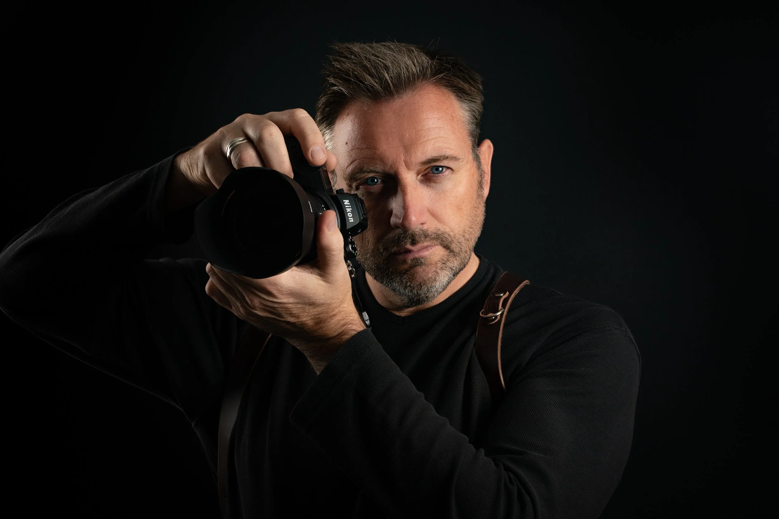 A man with short brown hair and a beard, wearing a black shirt, holding a professional camera up to his eye, against a dark background.