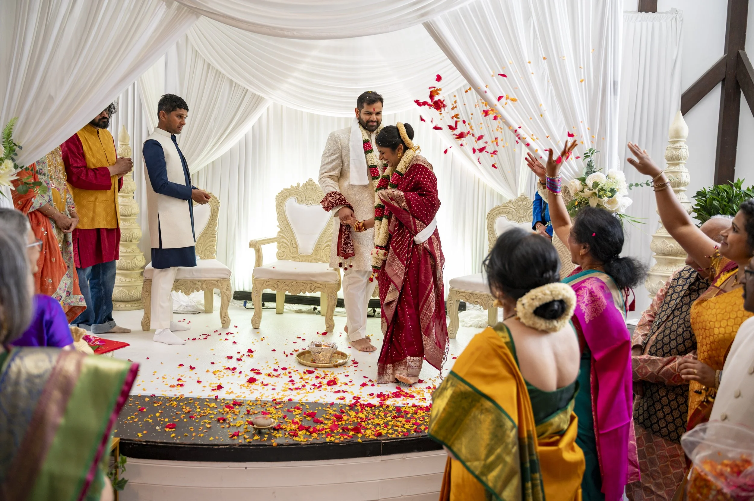 An Indian bride and groom celebrate on the stage whilst petals are thrown by guests