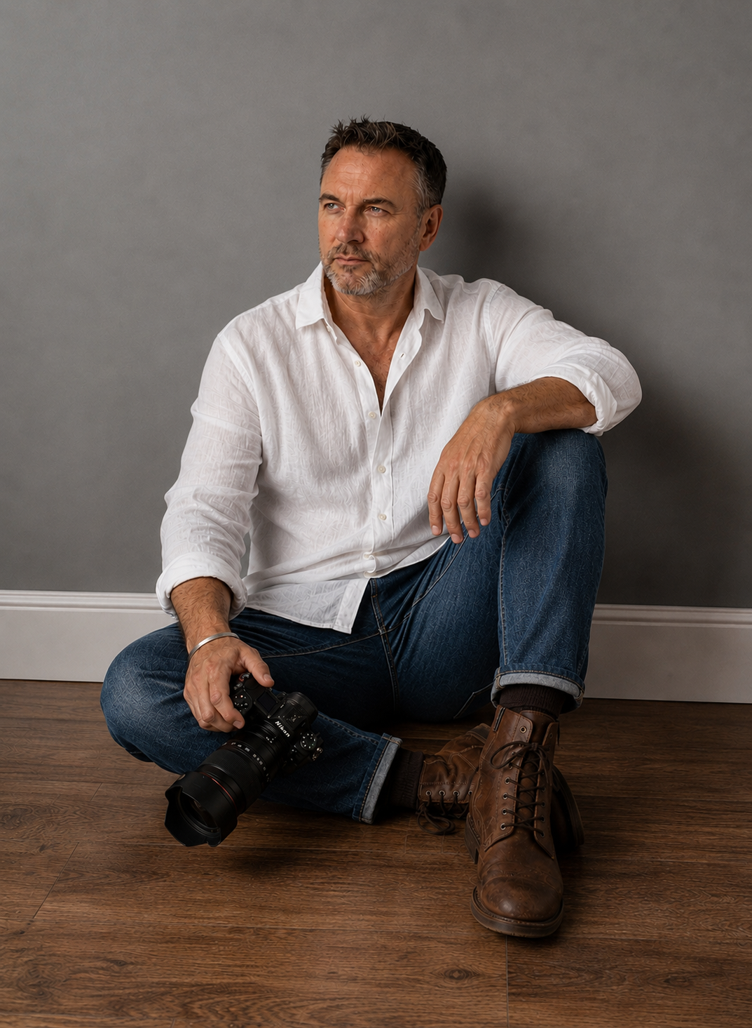 A middle-aged man with a grey beard and hair, wearing a white button-up shirt and blue jeans, sitting on the wooden floor against a gray wall. He has a camera in his right hand and is looking thoughtfully to his left.