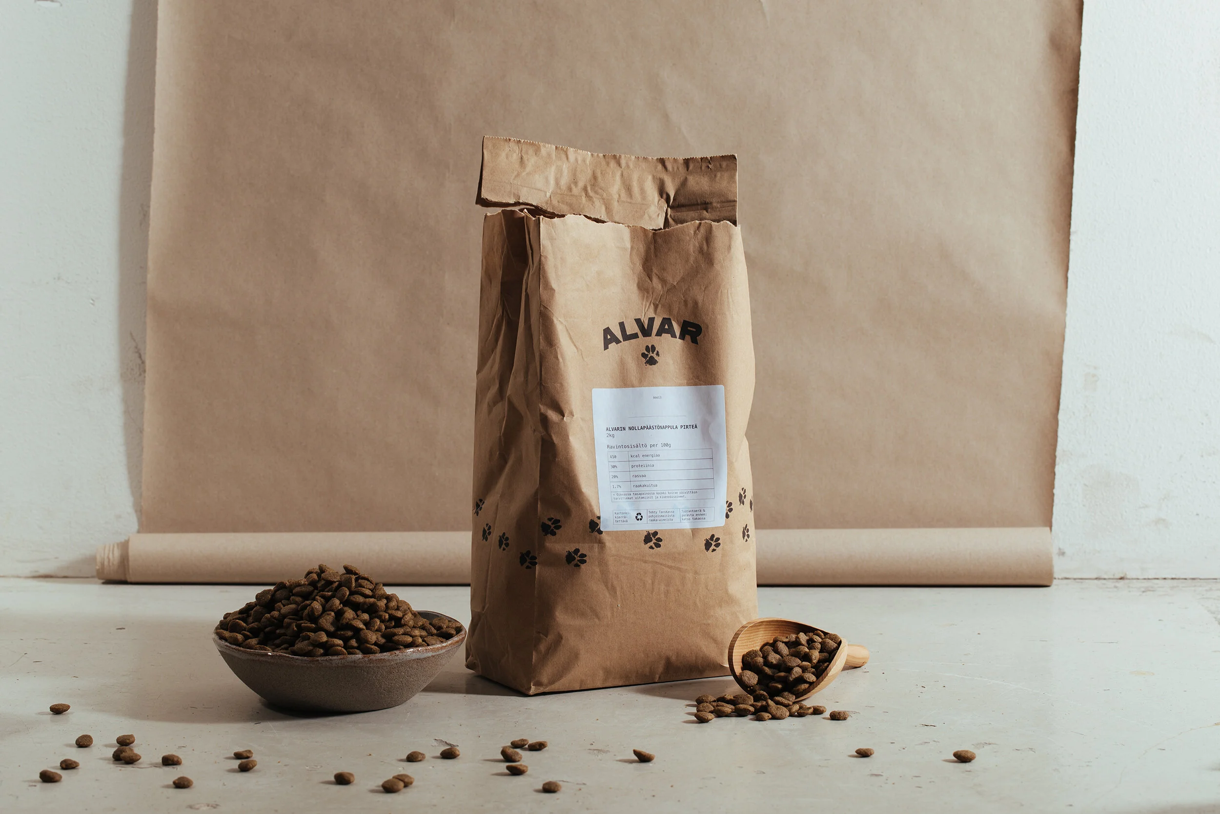 A brown paper bag of dog food labeled 'ALVAR' standing on a white surface with spilled kibble and a small bowl of kibble in front, against a background of a brown paper sheet and a white wall.