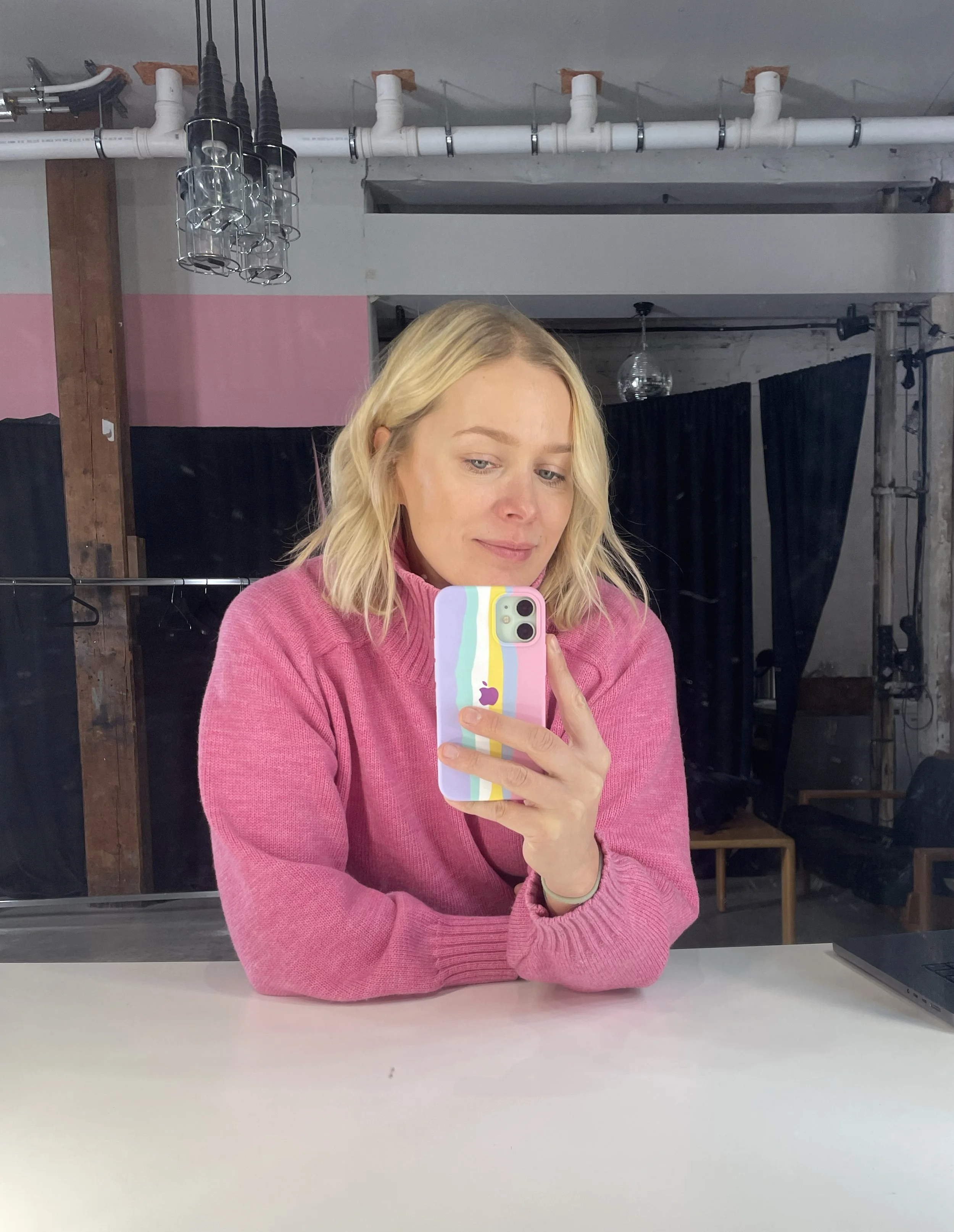 A woman with blonde hair wearing a pink sweater taking a mirror selfie with a colorful striped phone case in a studio or workspace with black curtains, wooden beams, and exposed pipes.