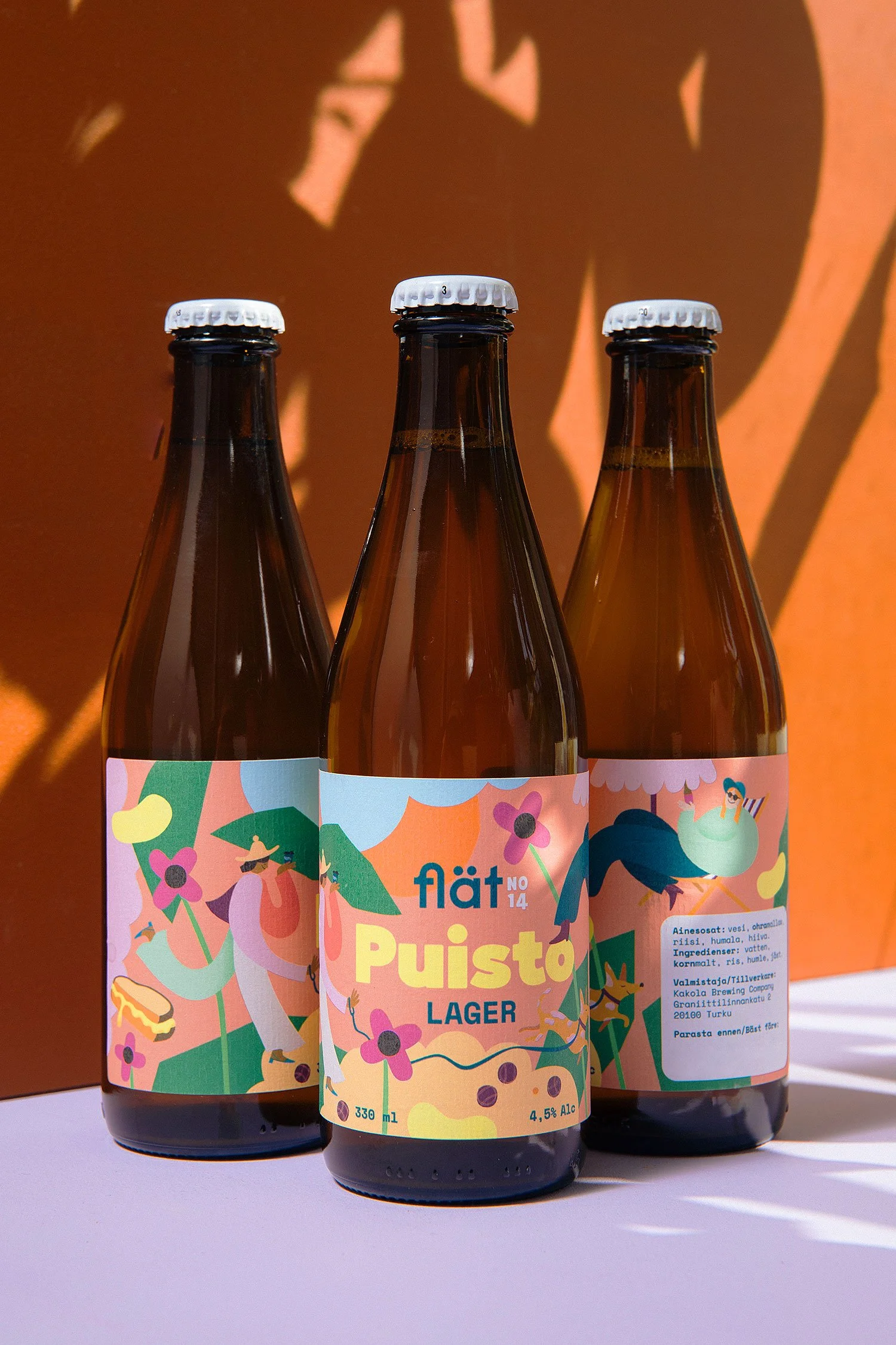 Three brown Bottles of beverage with colorful, tropical-themed labels featuring illustrations of people, toucans, and flowers, placed on a white surface with orange background and shadows.