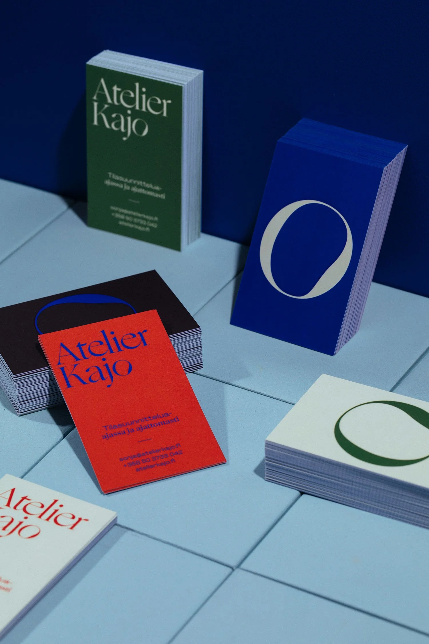 Several stacks of business cards or flyers on a table, with the words 'Atelier Kajo' and various text in Finnish printed on them, placed against a blue wall.