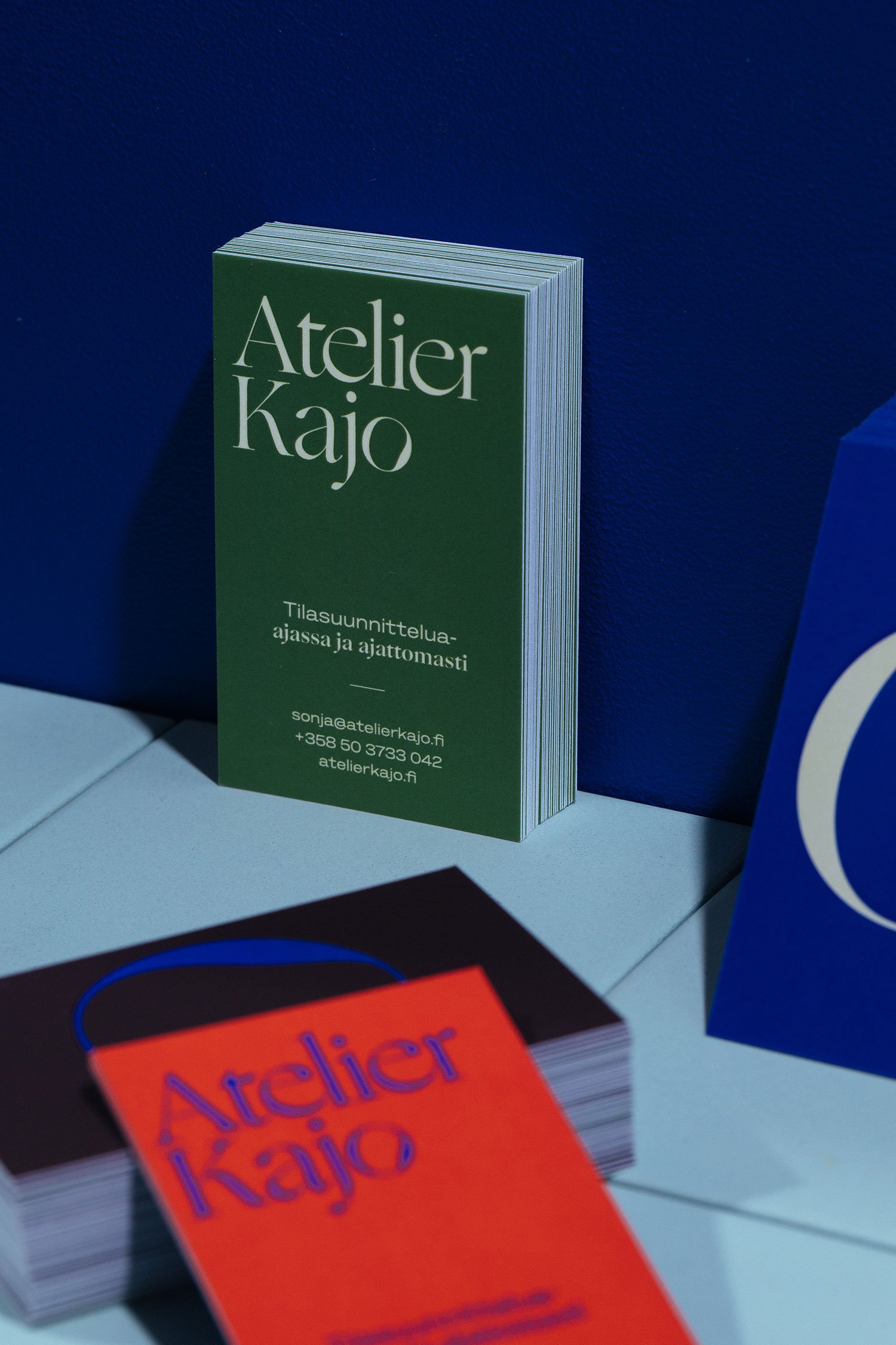 Close-up of a green business card for Atelier Kajo, which specializes in space planning, with contact details, set against a dark blue background with other blurred cards nearby.