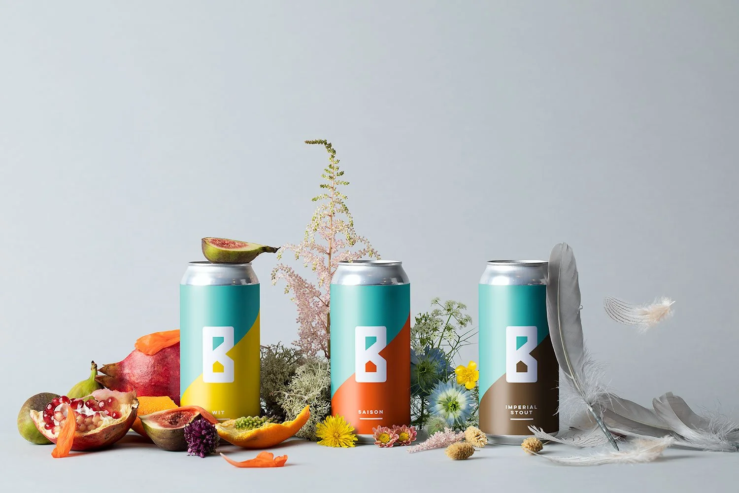 Three colorful cans of beer surrounded by assorted fruits, flowers, and feathers on a light gray background.