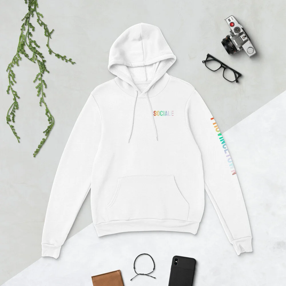 Flat lay of a white hoodie with "SOCIAL E" text, vintage camera, black glasses, plant, brown wallet, black bracelet, and smartphone on a gray background.