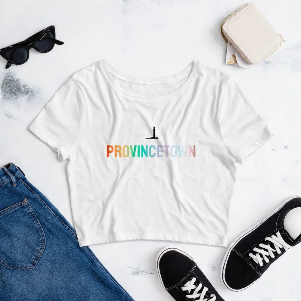 White t-shirt with "Provincetown" in rainbow colors and a lighthouse graphic, surrounded by sunglasses, jeans, sneakers, and a small wallet on a marble surface.