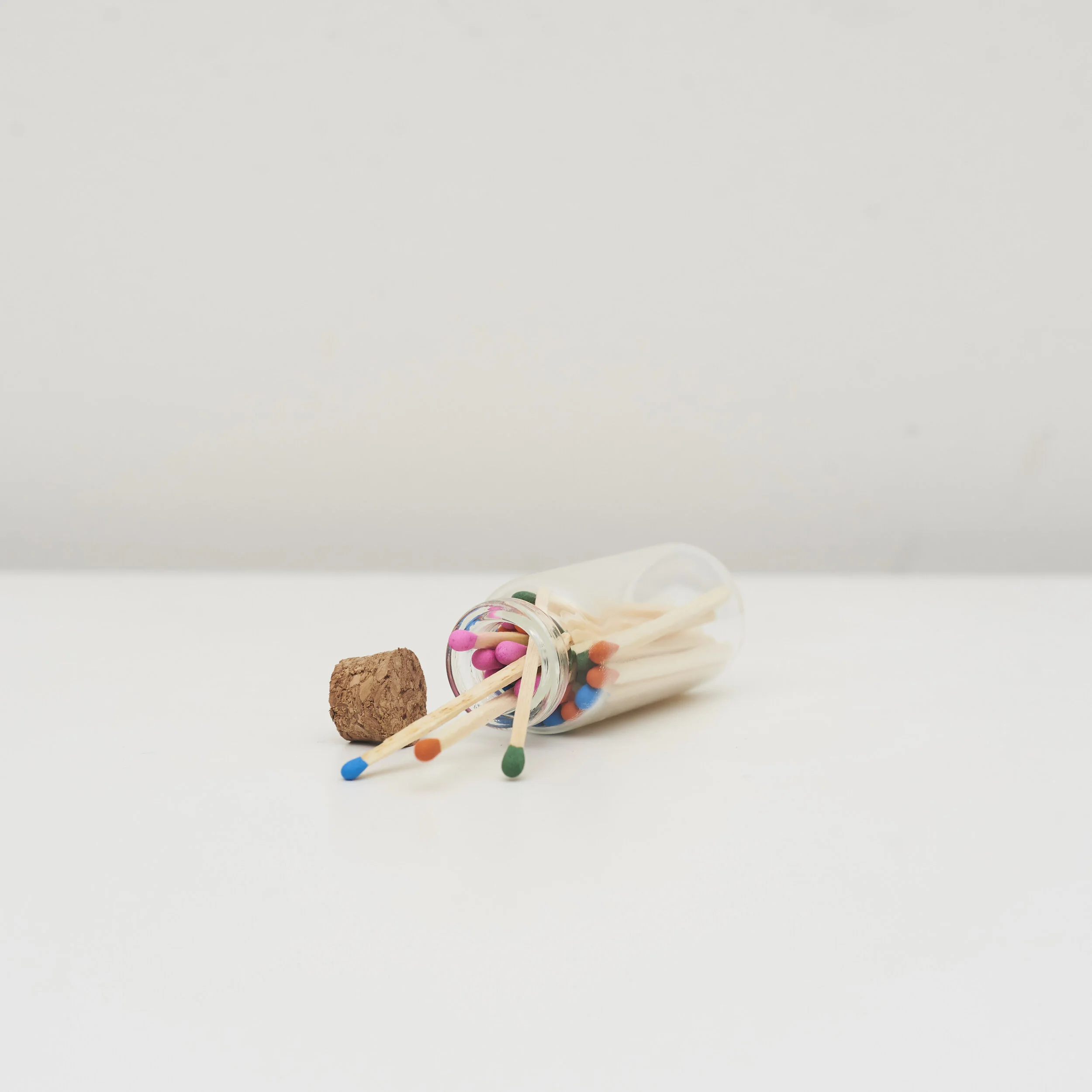 Colorful matchsticks in a glass vial with a cork stopper on a white surface.