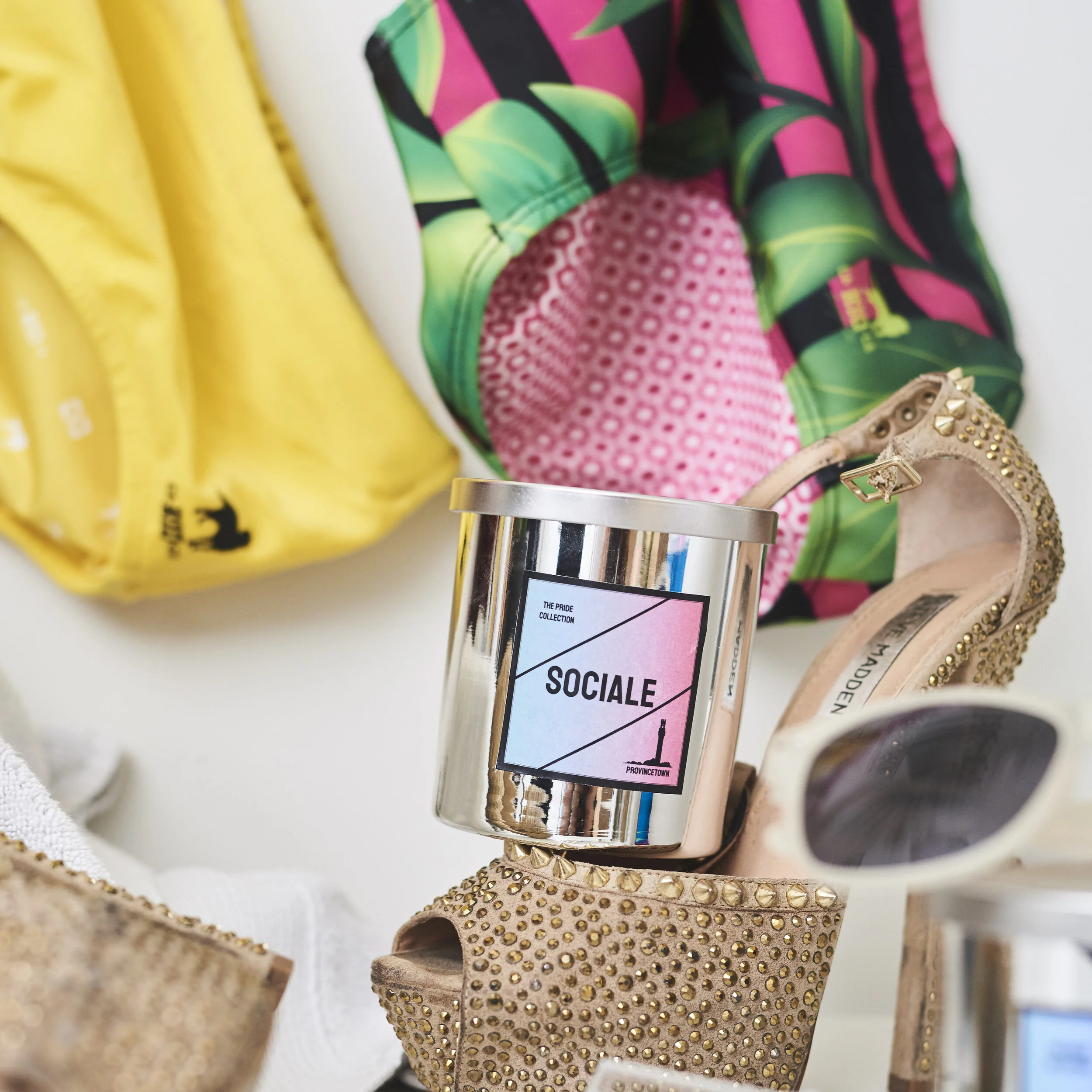 A silver candle labeled 'Sociale' is placed among gold studded high heels, sunglasses, a yellow garment, and a colorful patterned fabric in a close-up arrangement.