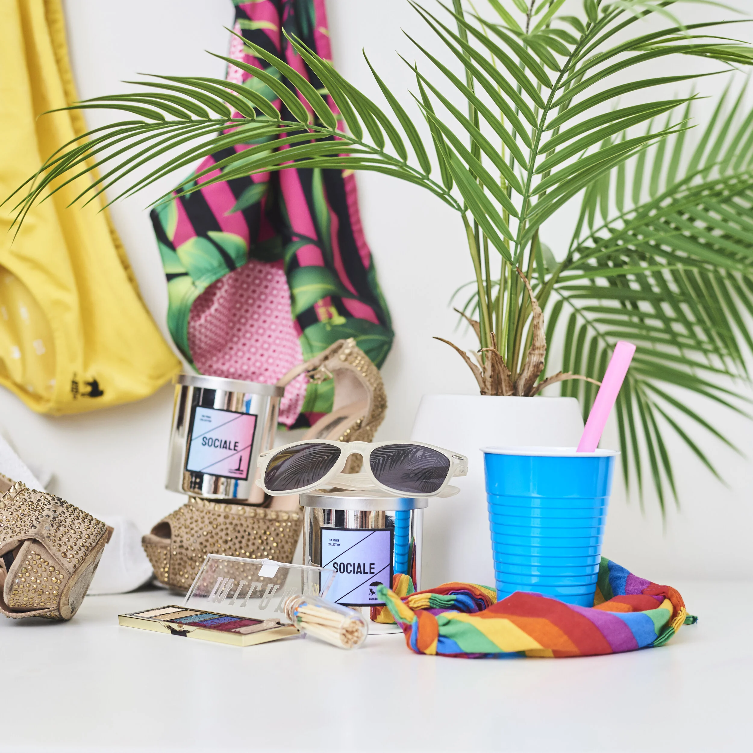 Summer-themed arrangement with a potted plant, blue plastic cup with pink straw, sunglasses, colorful scarf, makeup, gold high heels, and vibrant clothing.
