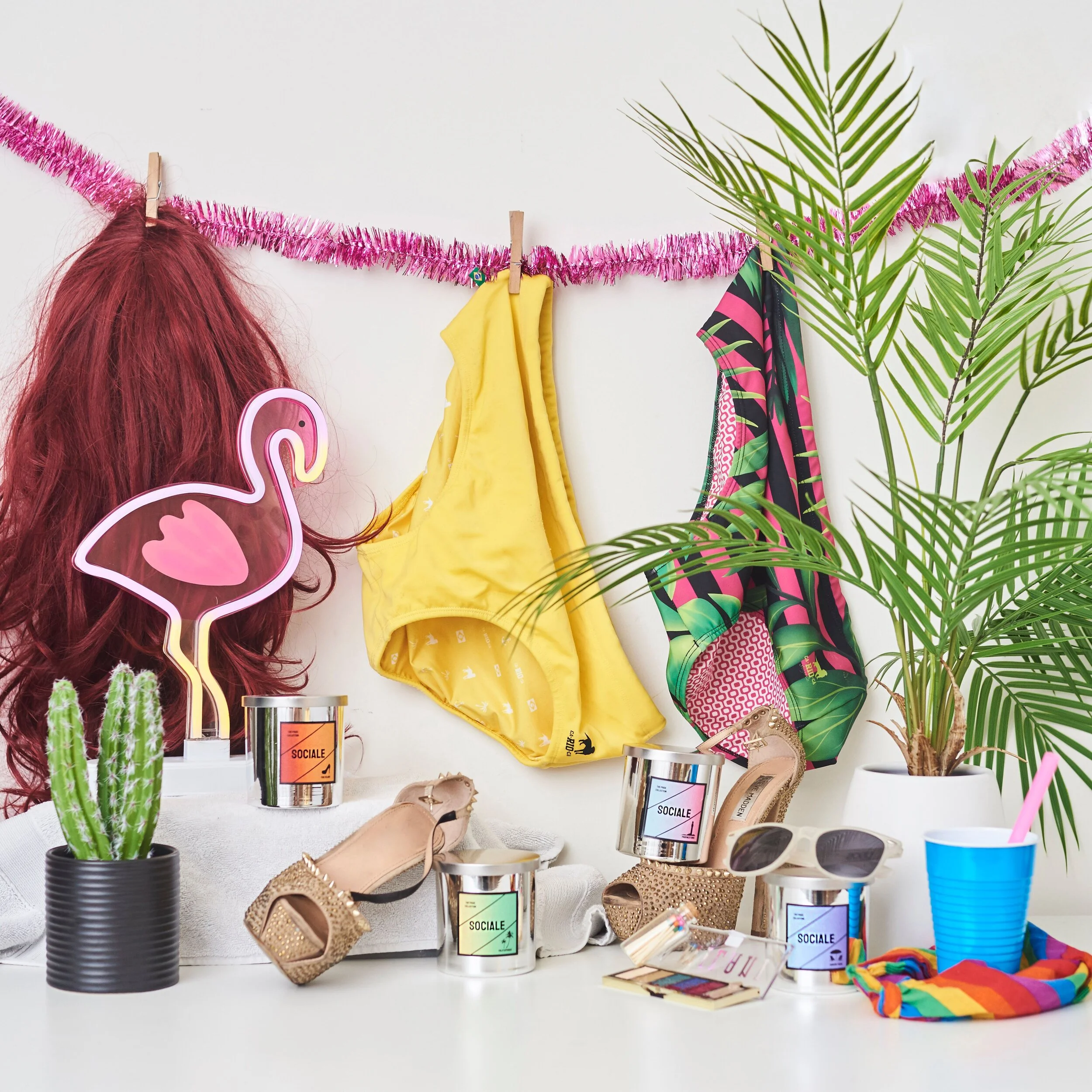 Table with decorative items including a pink flamingo light, a red wig, yellow and patterned clothing, candles, cactus and palm plants, high-heeled shoes, sunglasses, and a blue cup with a straw.