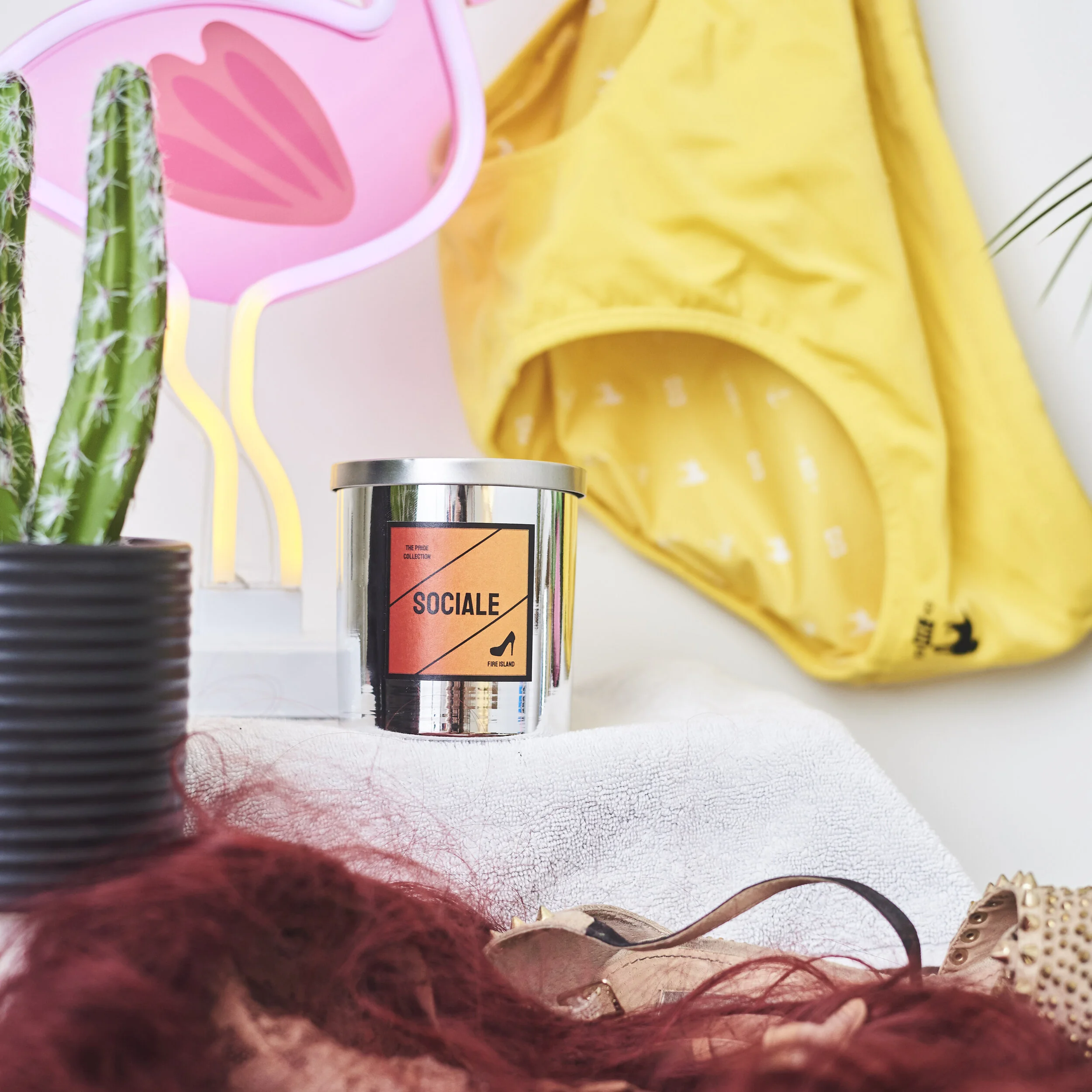 Decorative setup with a neon pink flamingo light, a small cactus in a black planter, a silver "Sociale" labeled candle, yellow shorts, and scattered accessories.