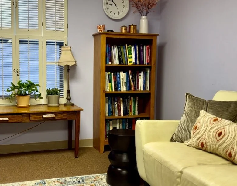 Living room corner with a wooden bookshelf filled with books, a yellow sofa with throw pillows, a side table, and a window with shutters.