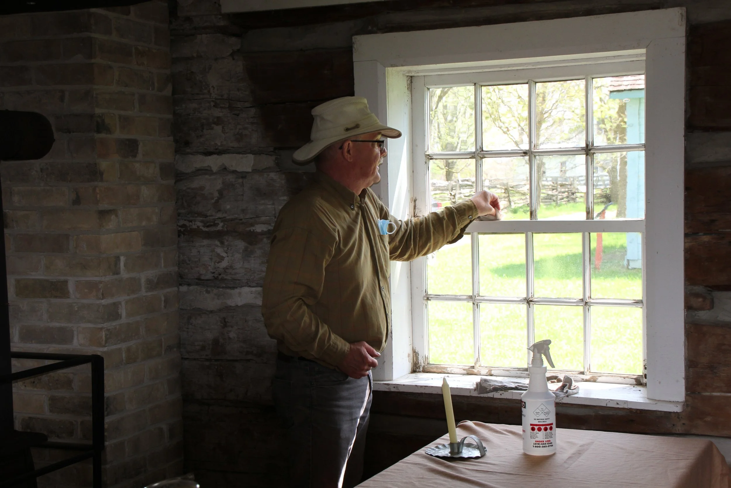 A man stands inside of a building, wiping window pains. A spray bottle sits on a table beside him.