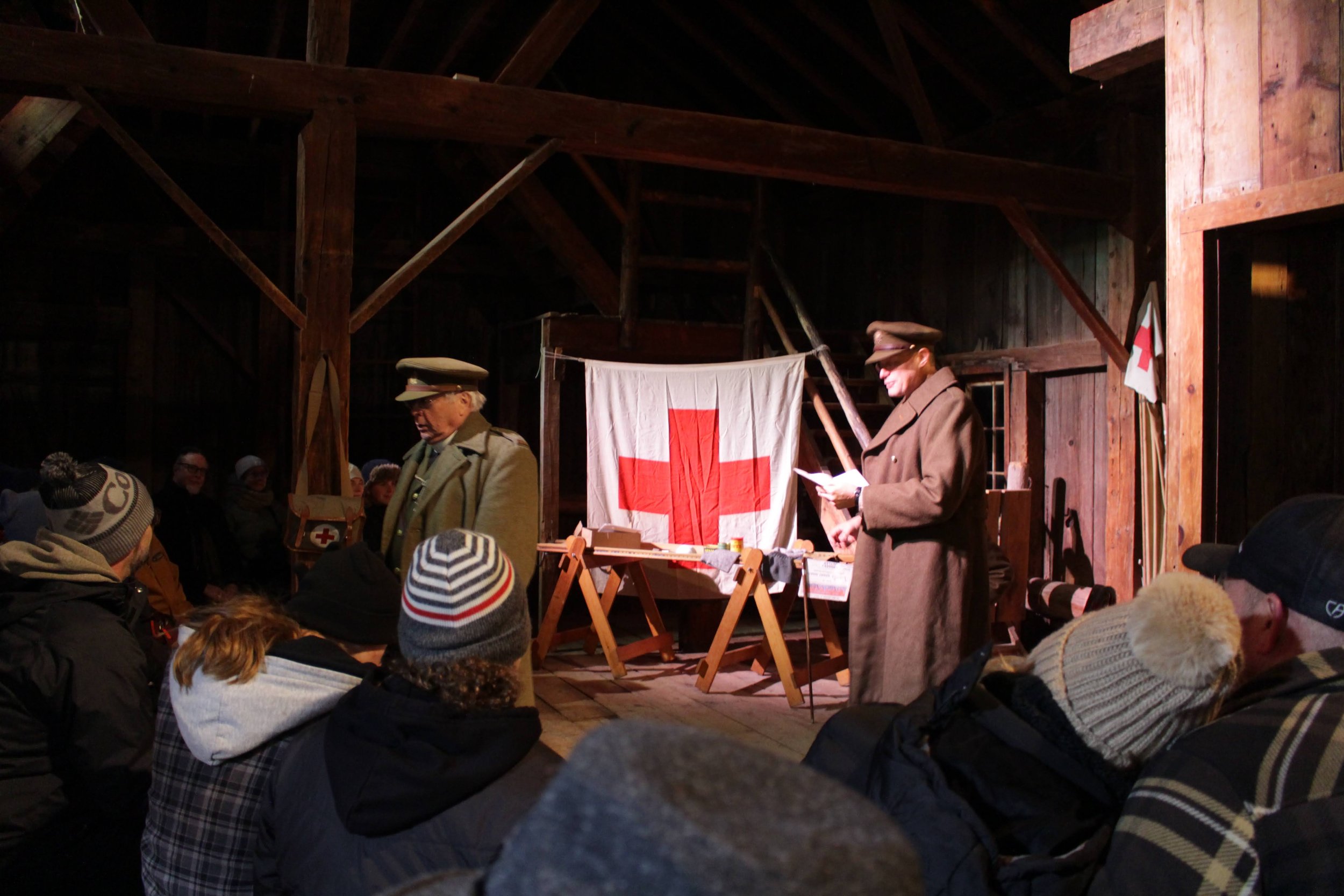 A photo of two World War 1 reenactors in officers' uniforms. They are playing out a scene inside a makeshift hospital. There is a large banner with a Red Cross symbol in the background.