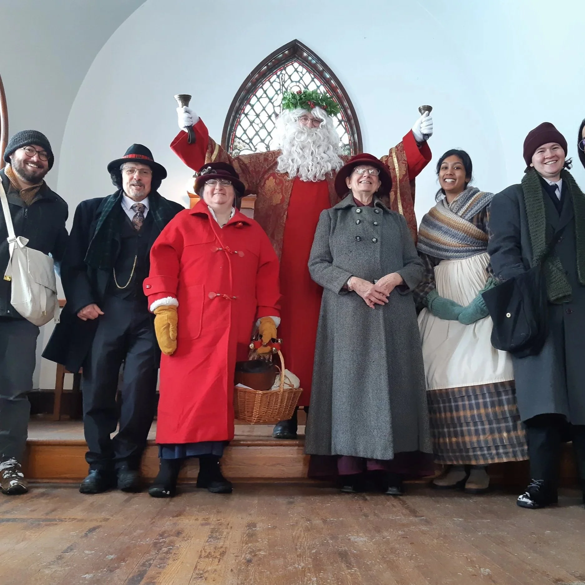 A photo of St. Nick surrounded by historical interpreters in 19th century dress. They are standing in the Trinity Anglican Church with a strained glass window behind them. St. Nick is holding up his bells.