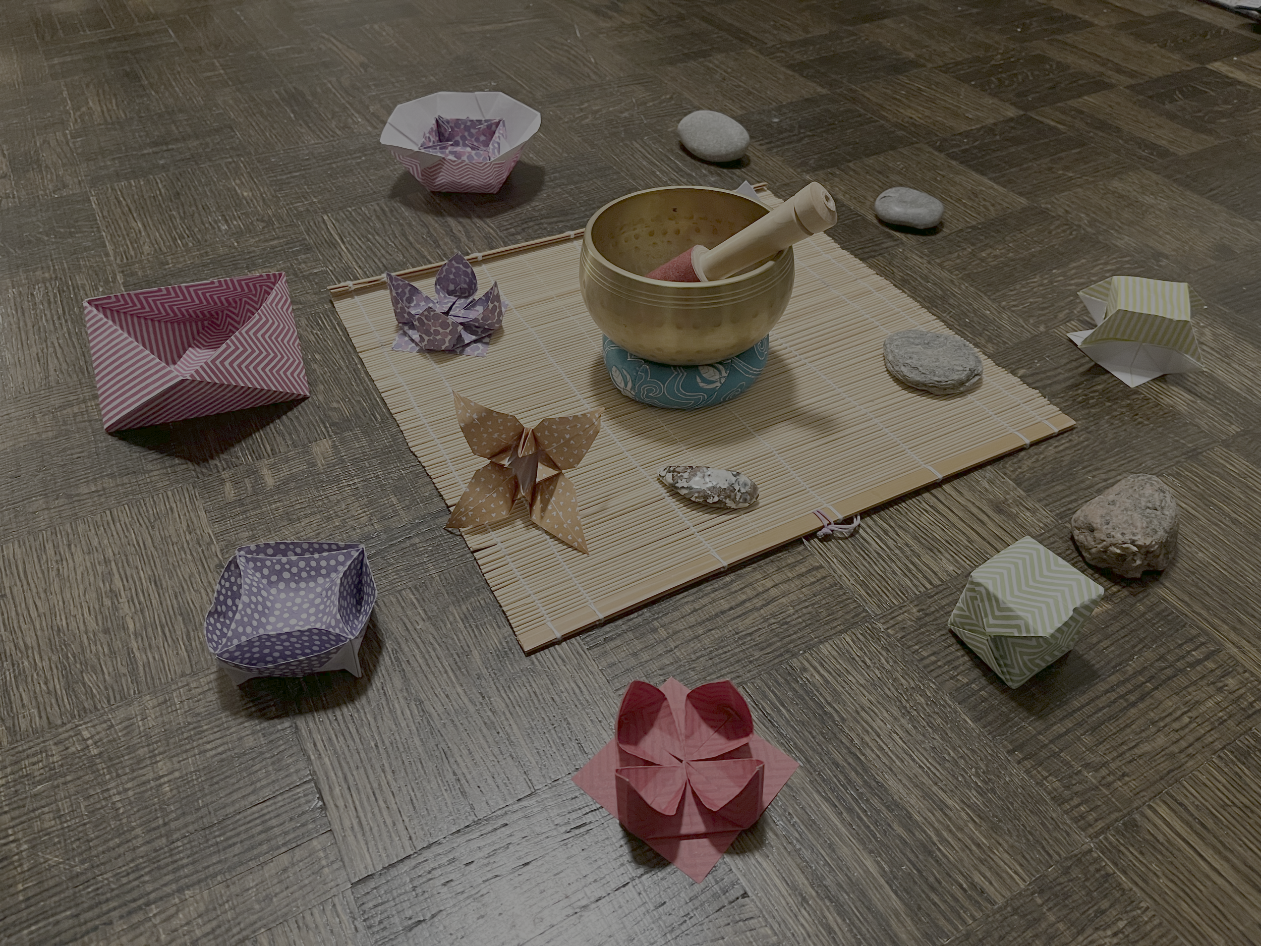 A ritual centerpiece created with colorful paper origami, a small brass bowl with a pestle inside, some stones, and a bamboo mat on a wooden floor.
