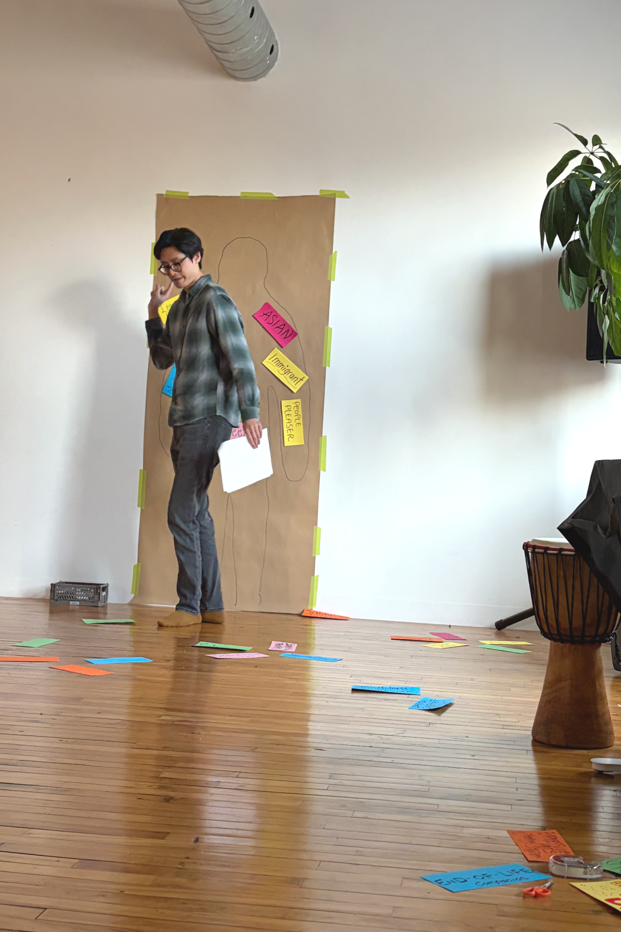 A person standing on a wooden floor with scattered colorful notes and papers, next to a large brown paper poster with various notes taped to it.
