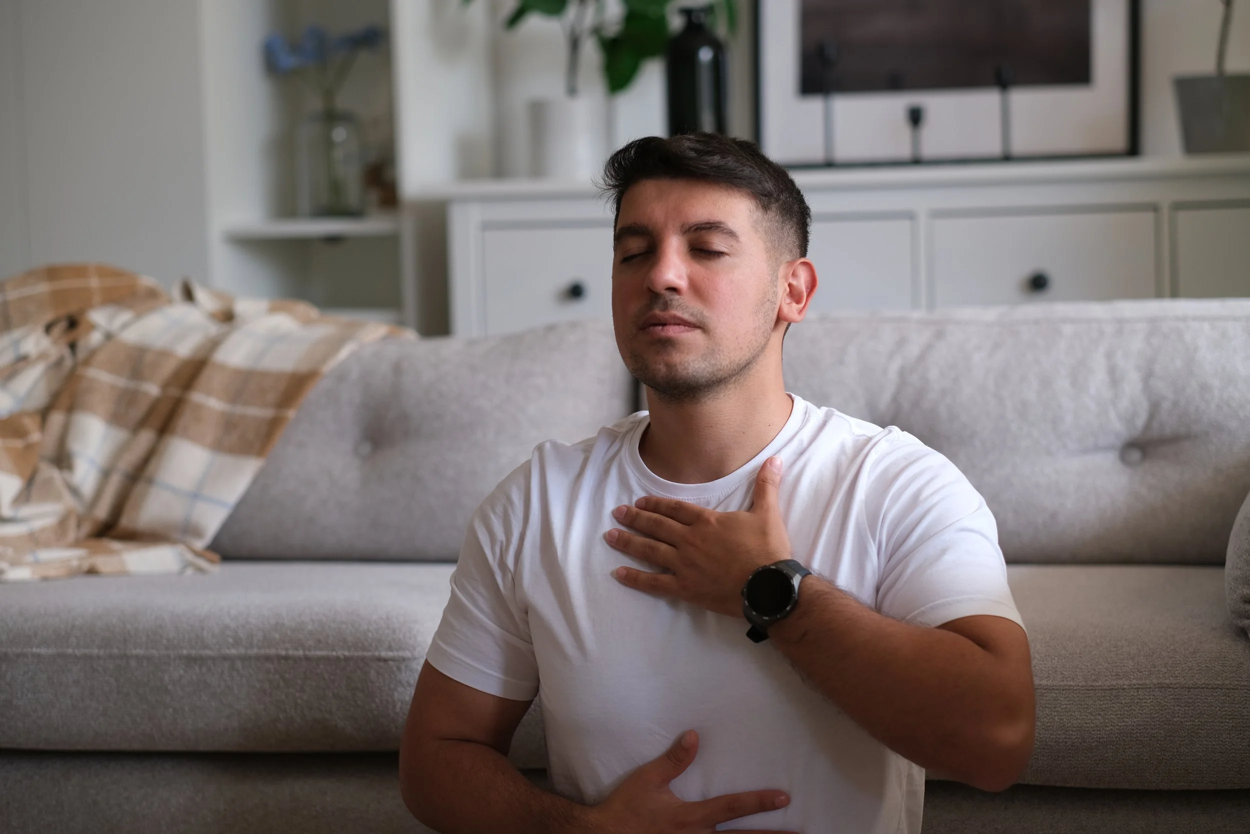 A young man with dark hair, wearing a white t-shirt and a black watch, sitting on the floor with his eyes closed, one hand on his chest and the other on his stomach, appearing to connect with his body and emotions.