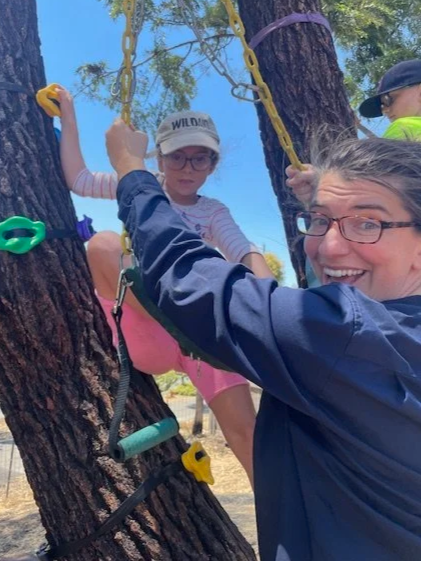 A woman and a young girl on a tree swing, with another person partially visible in the background, outdoors with trees and a clear blue sky.