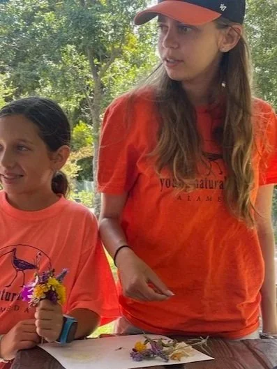 A person kneeling on grass, smiling, wearing a black hoodie and light-colored pants. Other people are partially visible in the background.