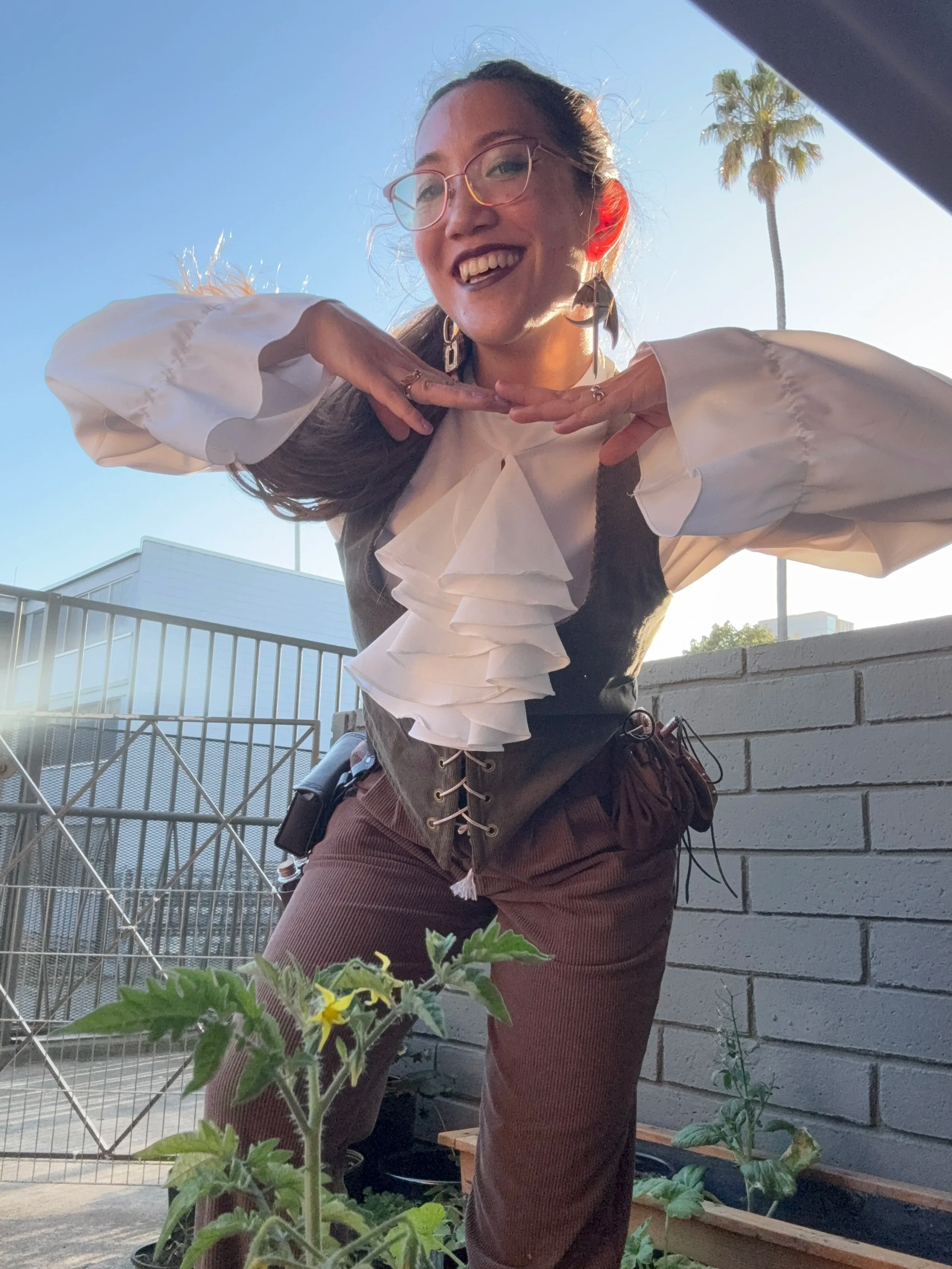 Woman with glasses and dark lipstick smiling and posing with hands under chin on a sunny rooftop garden, with palm trees and a blue sky in the background.