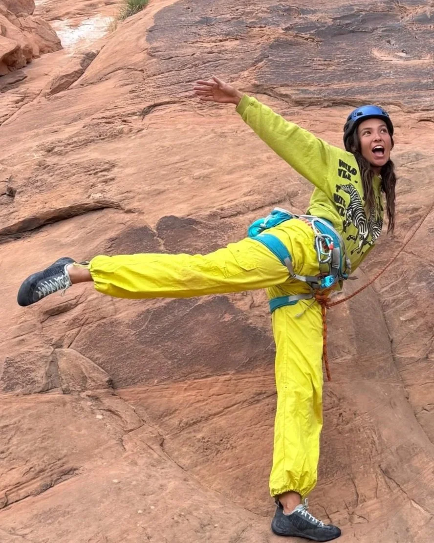 A woman rock climbing on a canyon wall, balancing on one leg with her other leg extended behind her and arms outstretched, smiling.