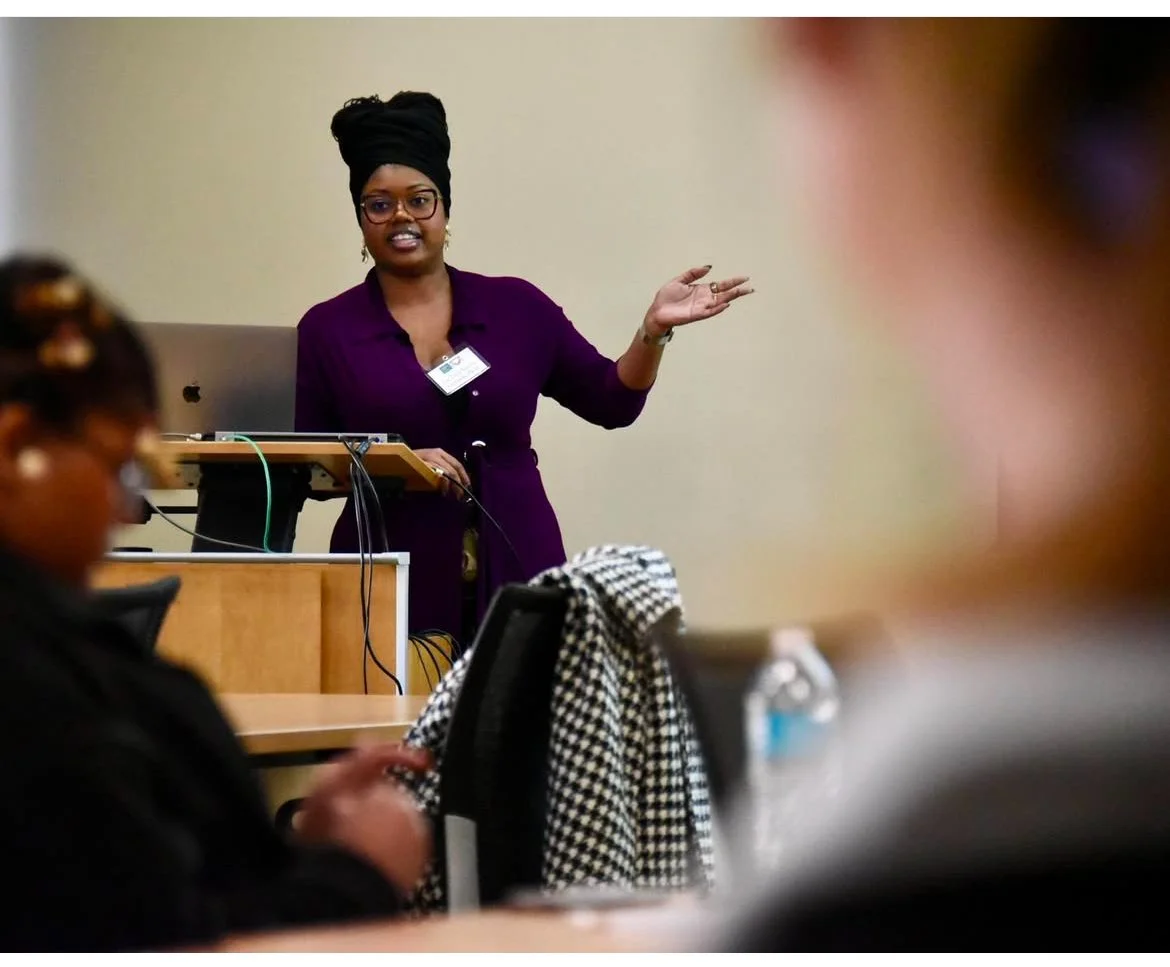 A woman with a black headwrap, glasses, and a purple shirt is giving a presentation in a classroom or conference room, standing near a podium with a microphone and a laptop. She is gesturing with her right hand while speaking.