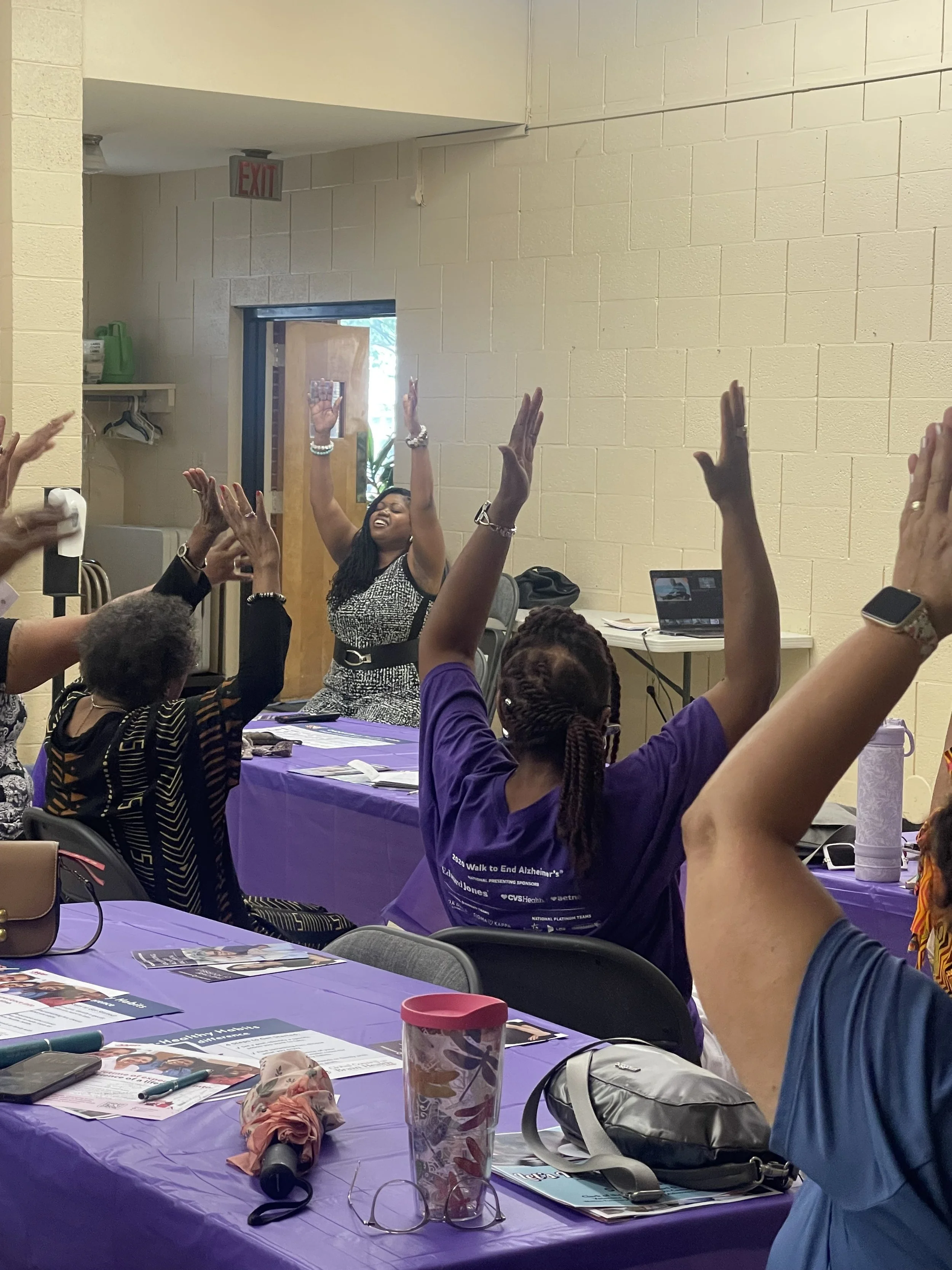 People sitting at tables with purple tablecloths participating in a meeting, with some raising their hands, and a woman at the front leading the activity, smiling and engaged.