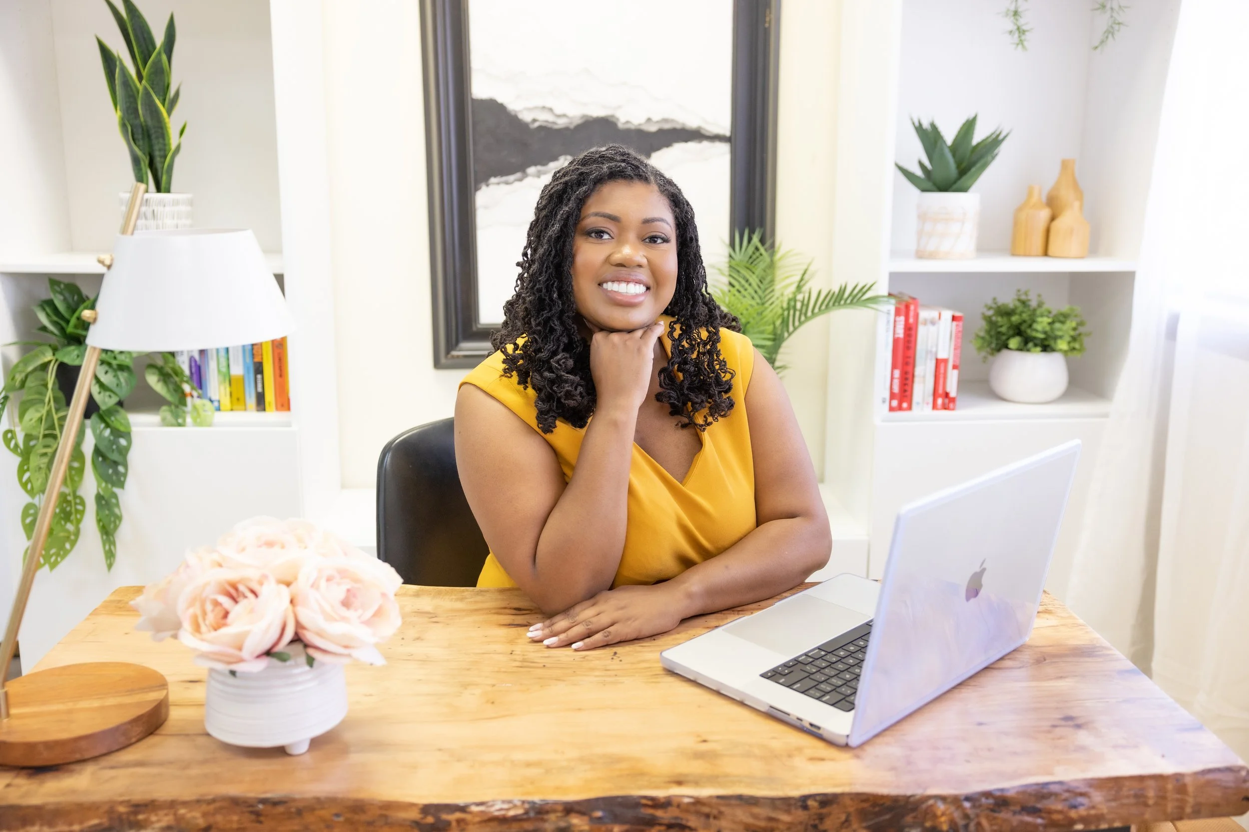 A woman with curly hair and a yellow dress sitting at a wooden desk with a laptop, smiling and resting her chin on her hand in a brightly decorated room with plants and books.