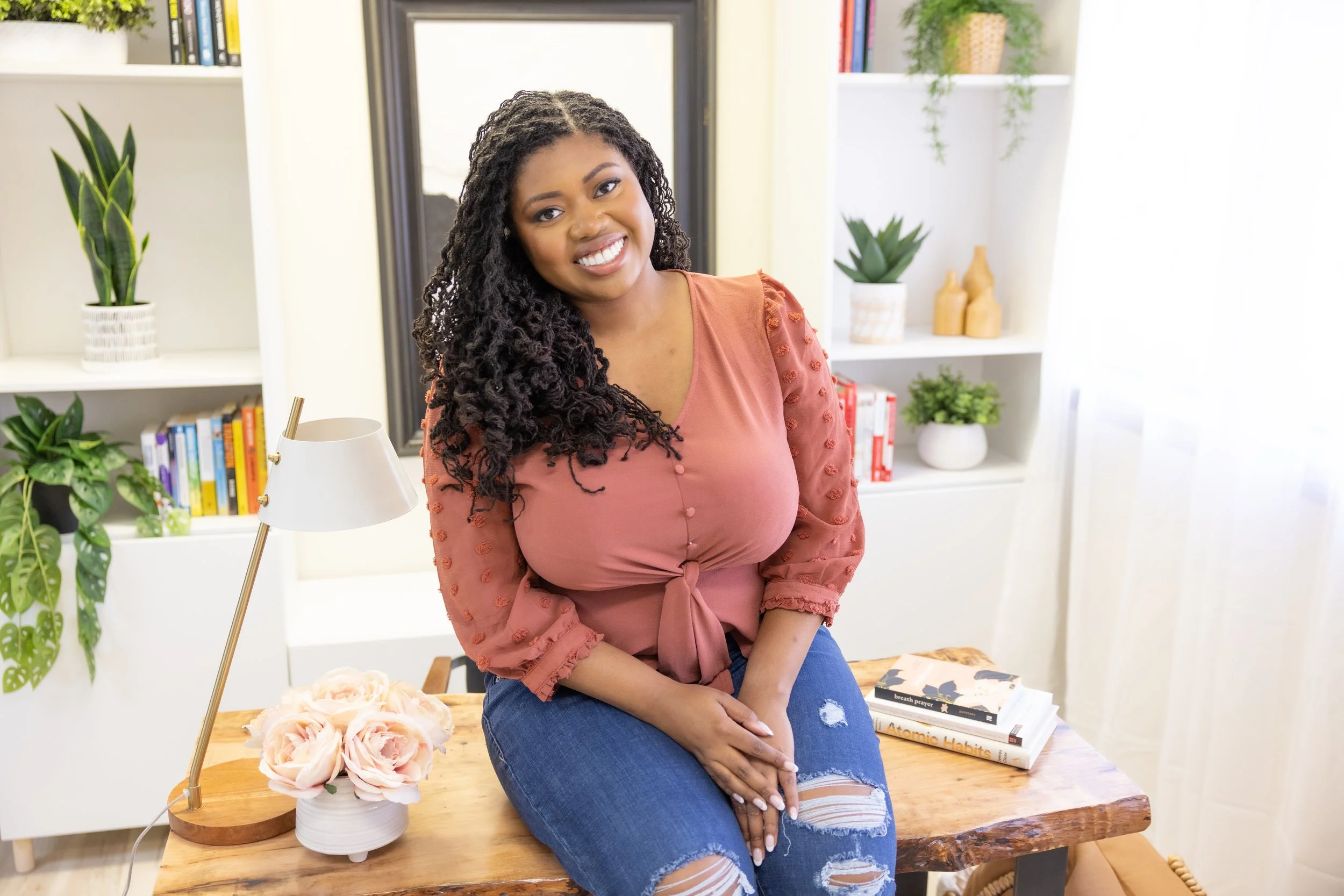 A smiling woman with curly hair wearing a pink blouse and ripped jeans sitting on a wooden table in a bright room with bookshelves and green plants.