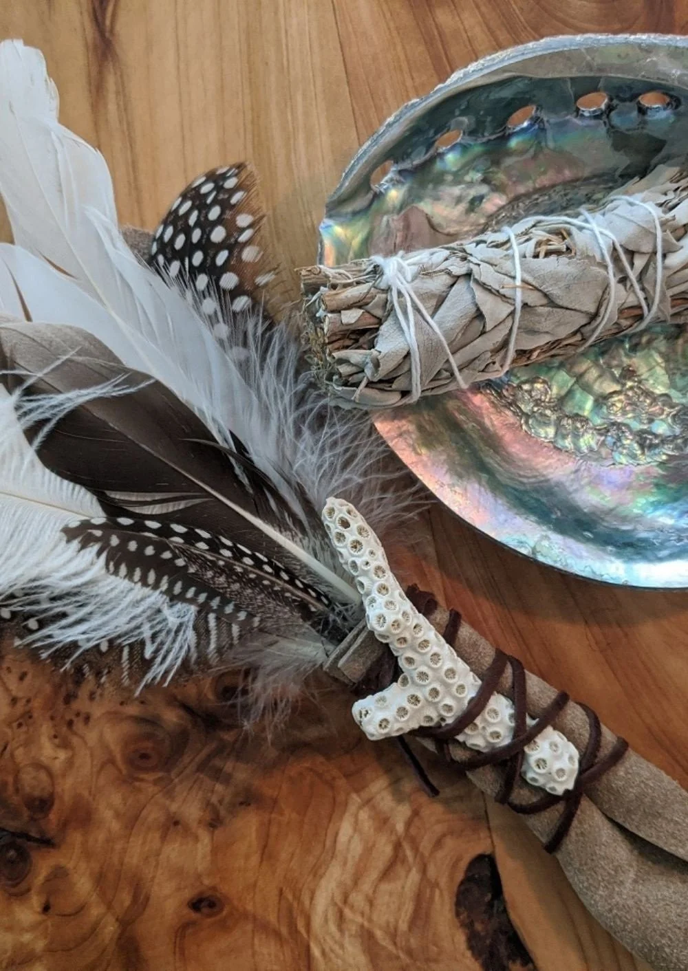 A woman lying face up on a massage table with a pink blanket, receiving a massage from another woman with curly black hair wearing a colorful shirt. They are in a room with a black wall, wooden chairs, and large green plants near a window.