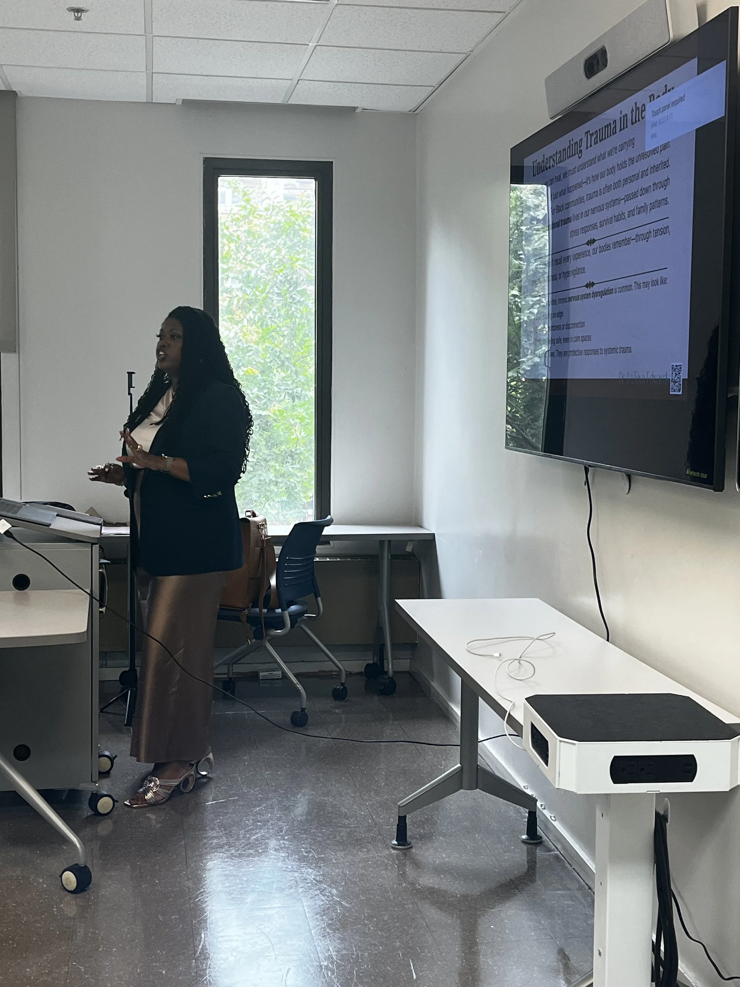 A woman giving a presentation in a conference room with a large electronic display screen, a window, and office chairs.