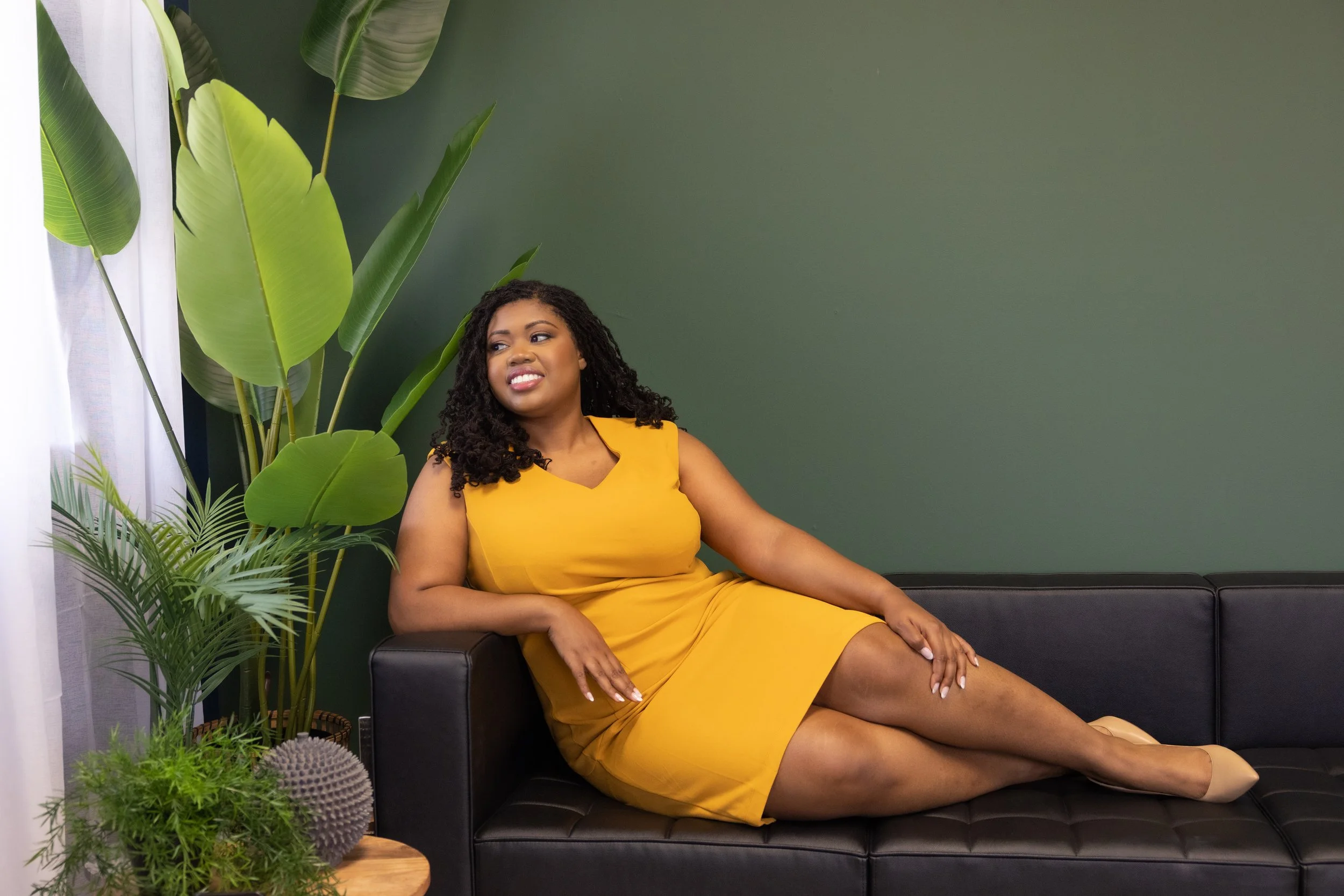 Smiling woman in a yellow dress reclining on a black sofa beside lush green houseplants