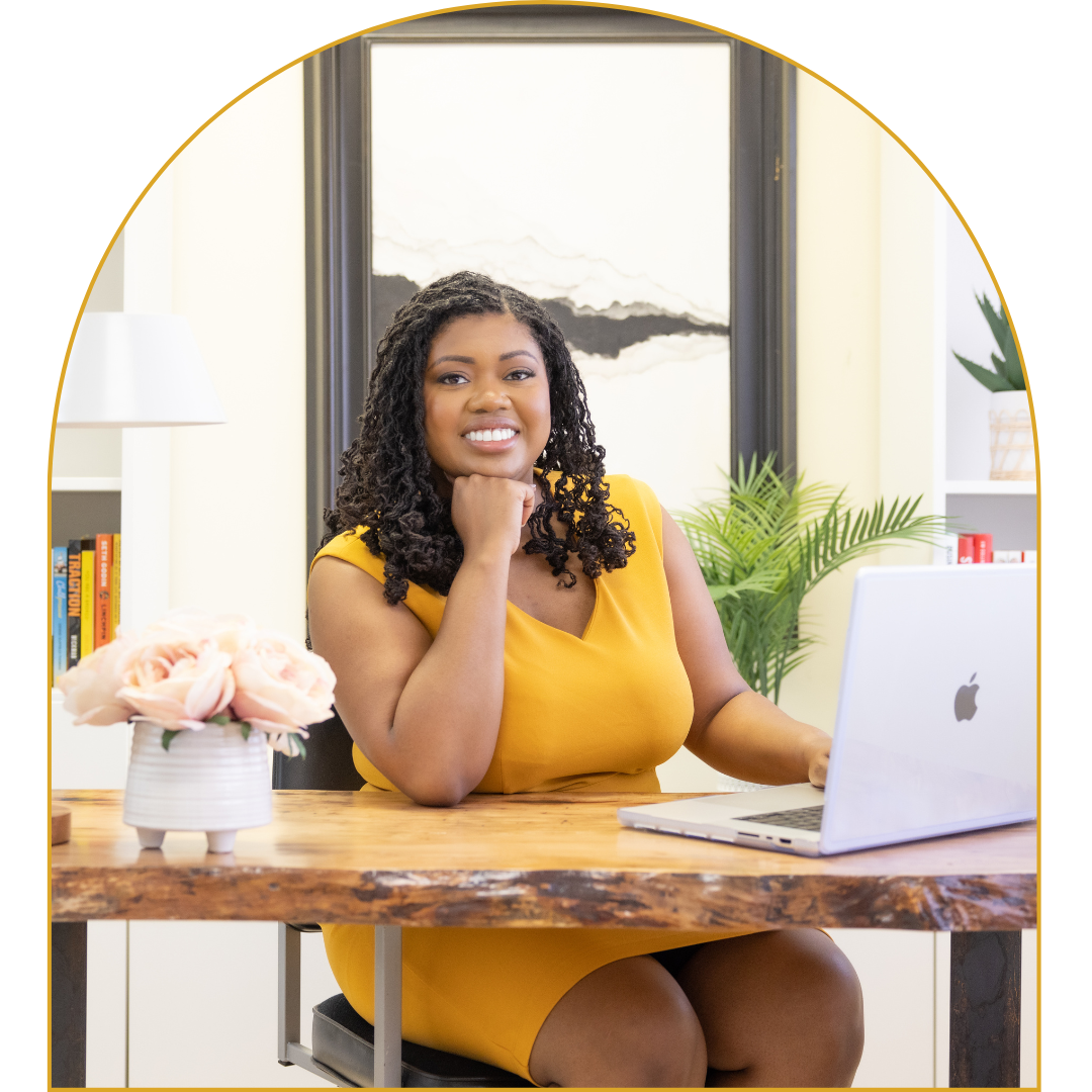 Woman in yellow dress sitting at wooden desk with laptop and pink roses in a vase, smiling, in a home office with plants and bookshelf.