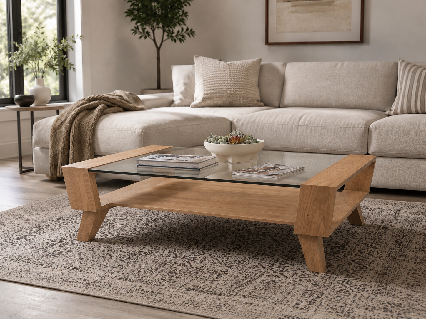 Living room with a beige sectional sofa, throw pillows, a wooden and glass coffee table with magazines and a succulent centerpiece, a patterned rug, a window with plants outside, and a framed artwork on the wall.