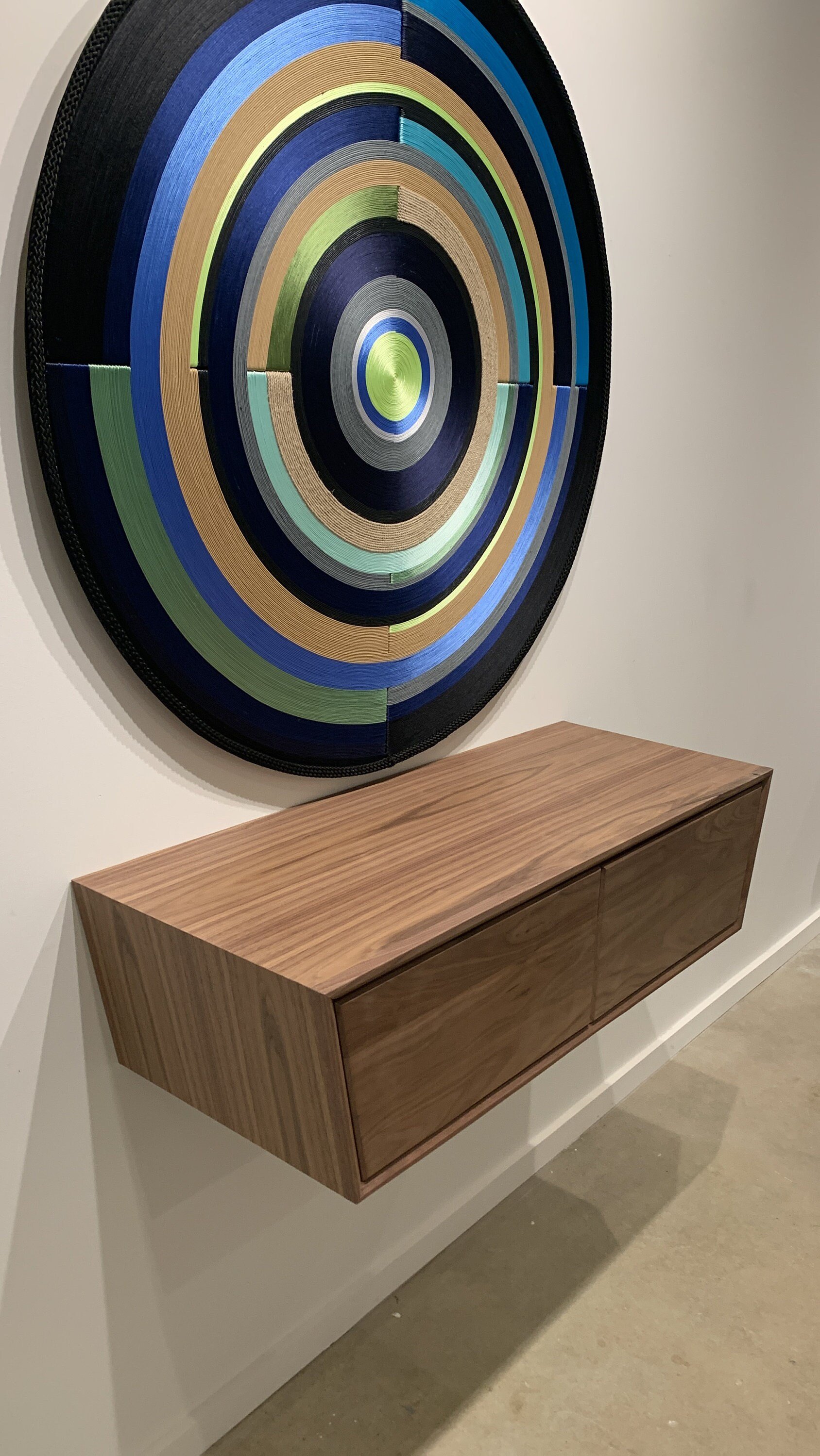 A circular artwork composed of concentric rings made of fabric in multiple colors, mounted on a white wall above a wooden sideboard.