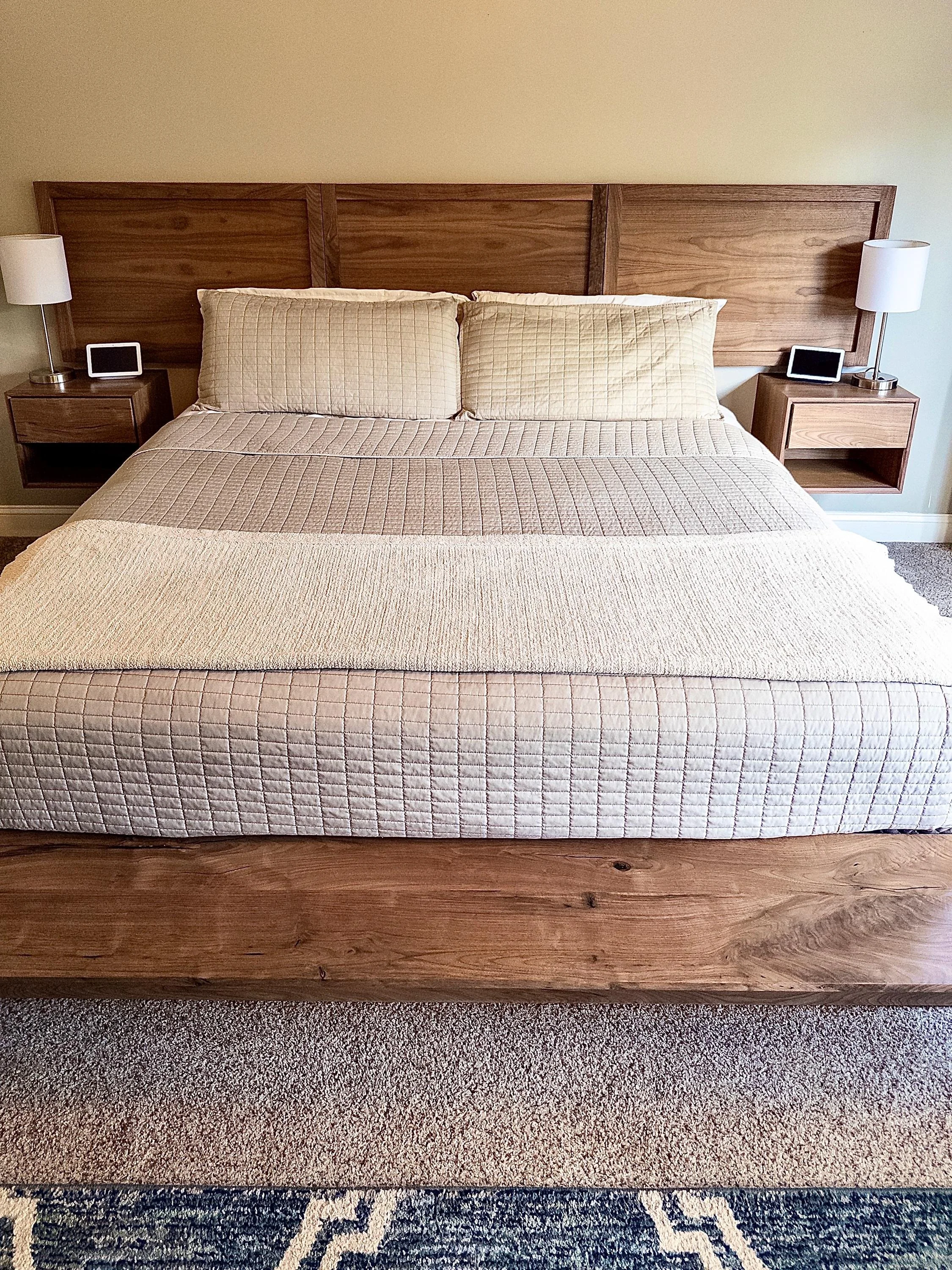 A bed with a textured beige bedspread and matching pillows, a wooden headboard, two bedside tables with lamps, digital clocks, and a patterned rug partially visible at the bottom.