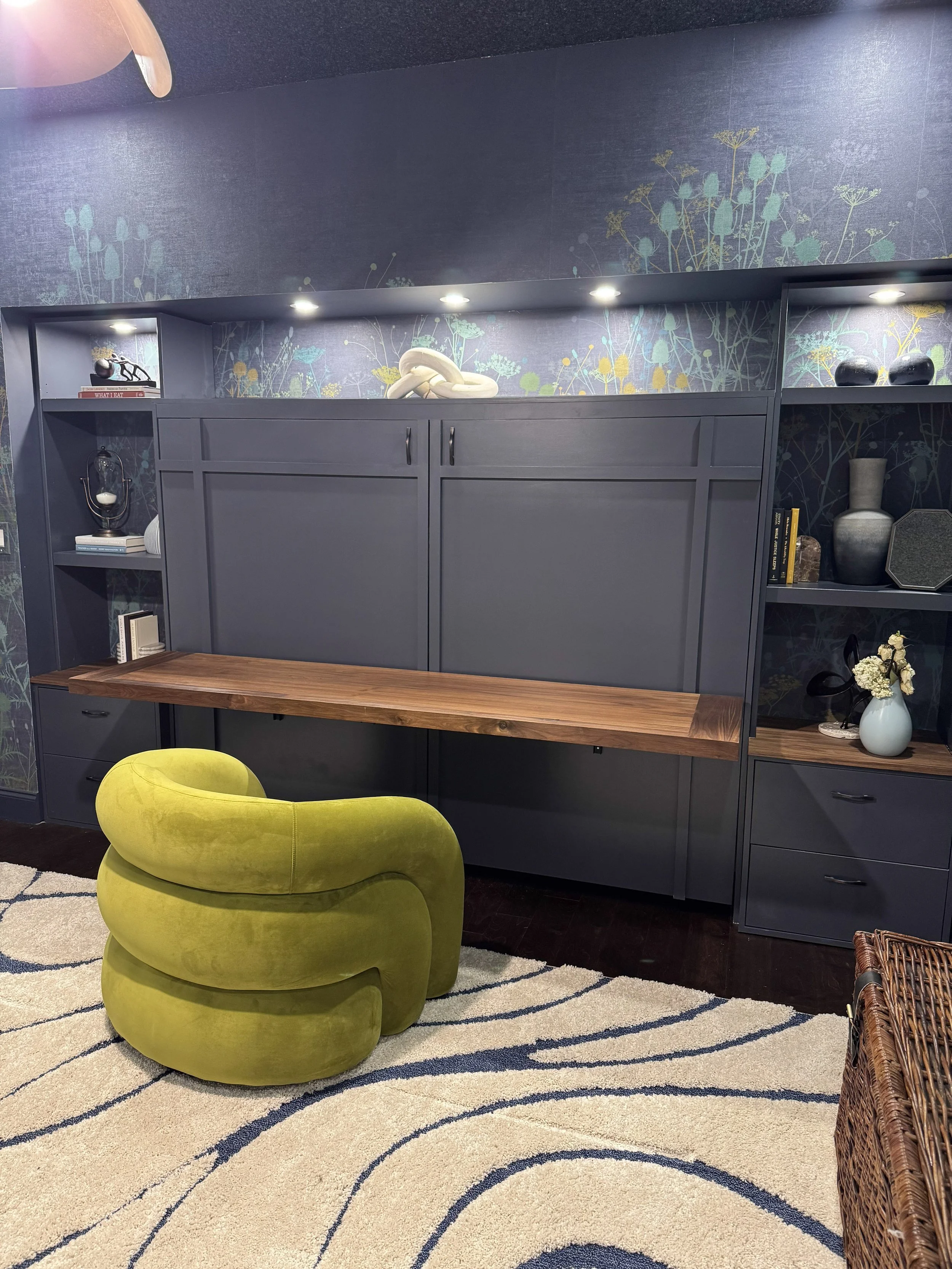 Room corner with dark blue built-in shelving unit, decorative items including books, vases, and sculptures, a yellow velvet chair, a patterned area rug, and dark hardwood flooring, against a blue wall with floral wallpaper.