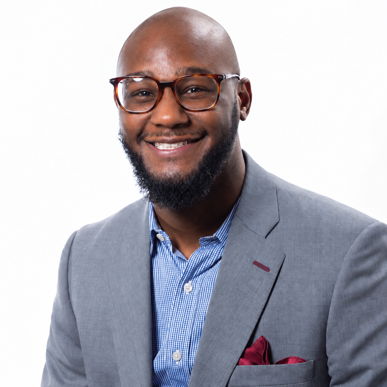 Portrait of a smiling African-American man with a beard, wearing glasses, a light gray suit, and a blue checkered shirt, against a white background.