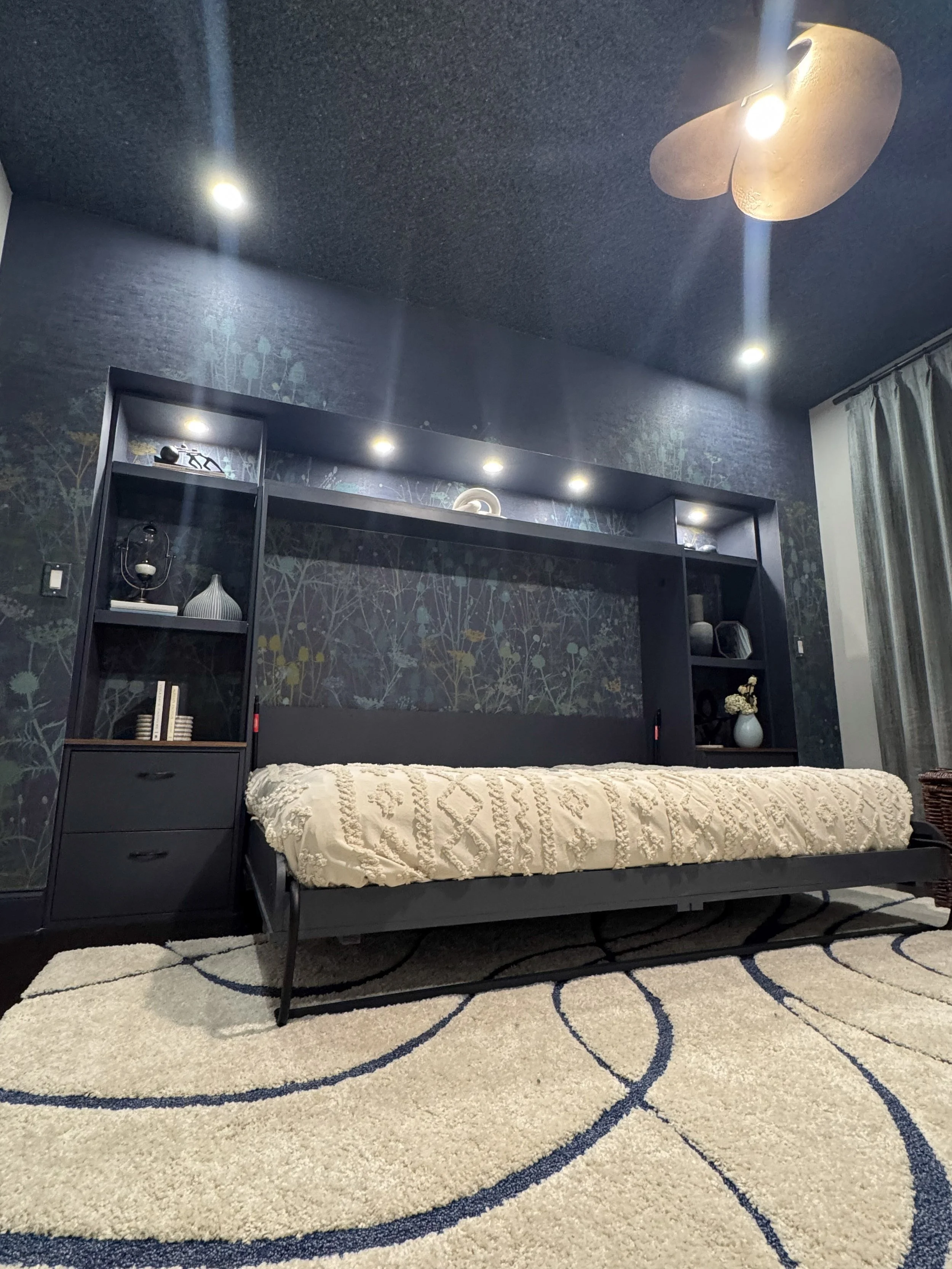 A bedroom with a black bed frame and a cream textured bedspread, a black bookshelf with decorative items, a wall with botanical wallpaper, a ceiling fan, and a beige rug with navy blue curved patterns.
