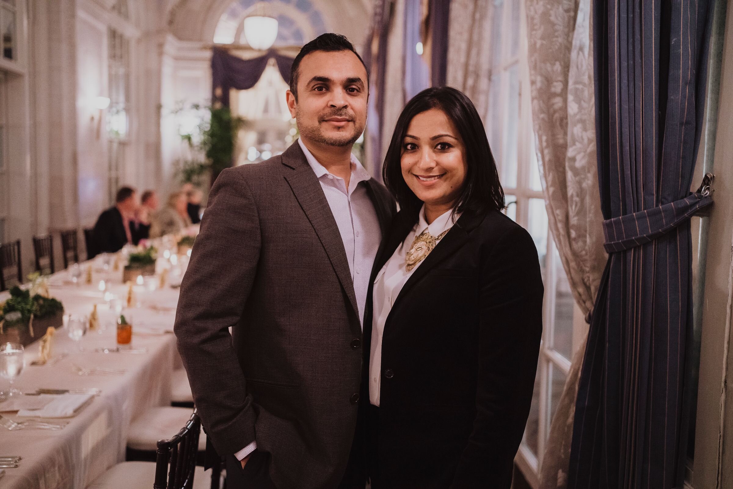 A couple in formal attire at an elegant indoor event with a long, decorated dining table in the background.