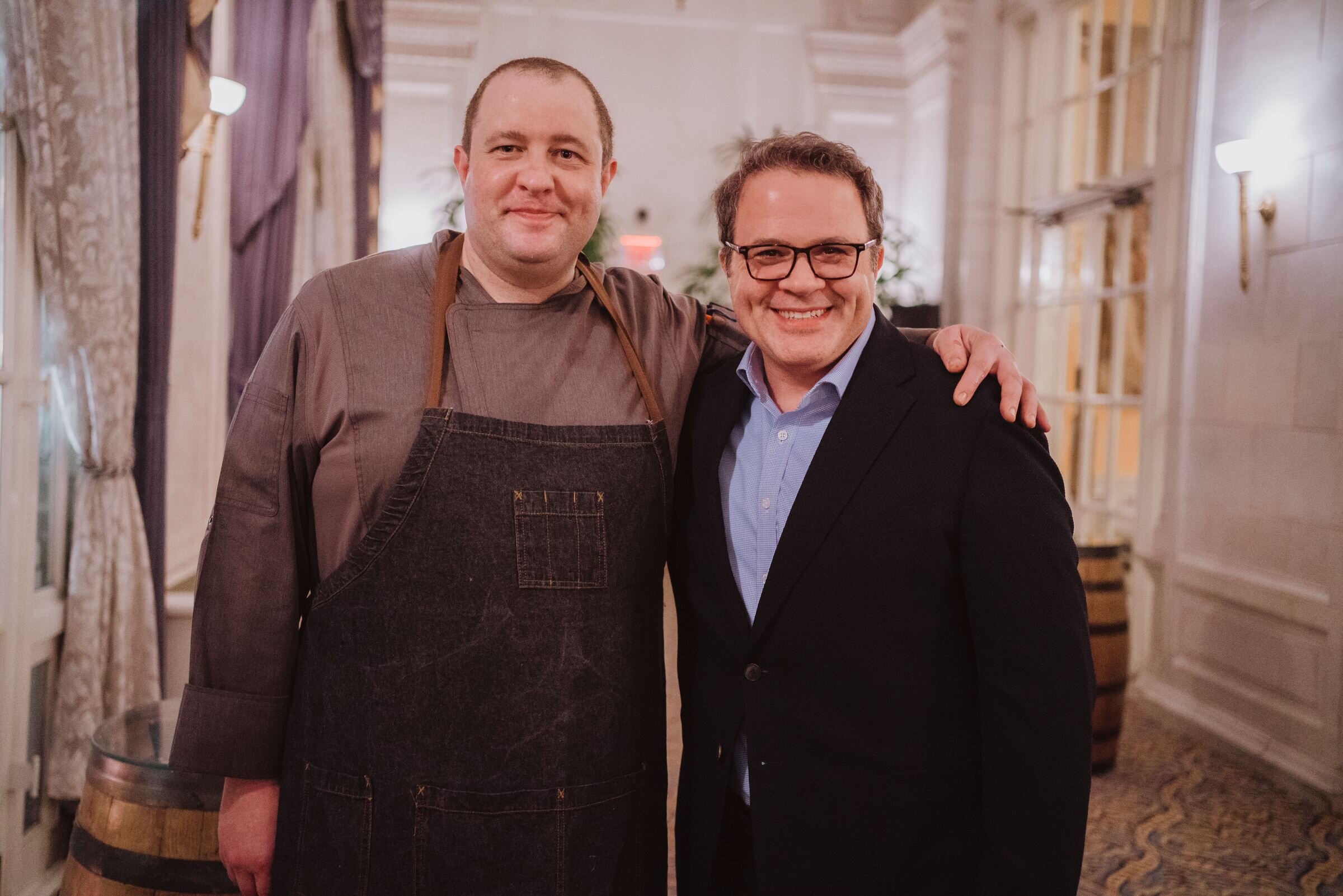 Two men standing together indoors, one wearing a chef's apron and the other in a suit, smiling at the camera.