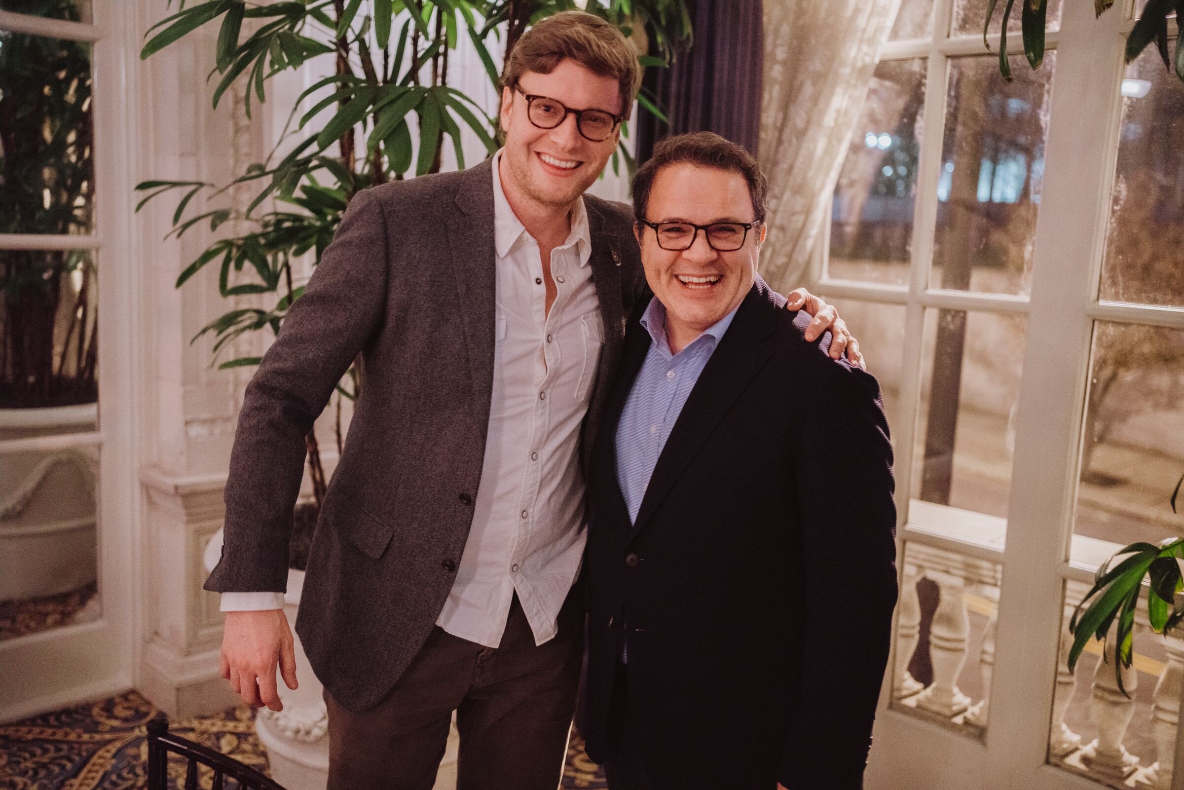 Two people posing together indoors, both wearing glasses and suits, standing in front of a large window, with plants in the background.