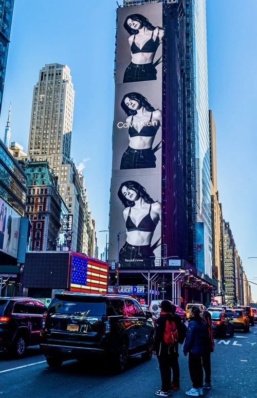 Street scene in Times Square, New York City, featuring a tall building with a large black-and-white Calvin Klein billboard displaying a model, surrounded by traffic, skyscrapers, and an American flag electronic sign.