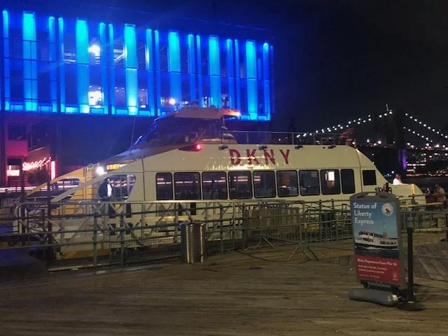 A ferry labeled "DKNY" docked at night, with a brightly lit building displaying blue lights in the background. There is a sign for the Statue of Liberty Express nearby. A bridge is partially visible in the distance.