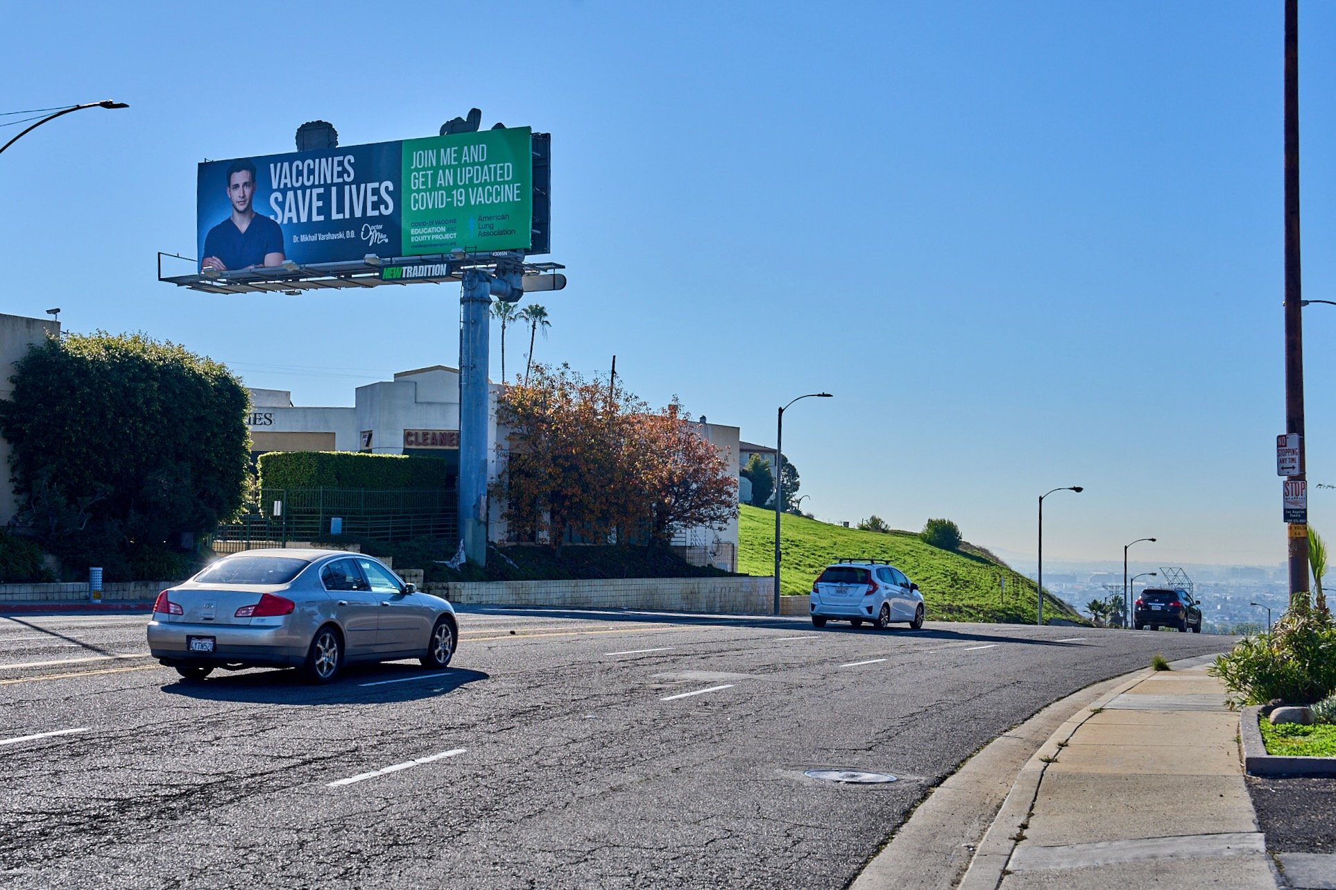 Street scene with a billboard promoting COVID-19 vaccine, featuring a doctor’s image and the slogan 'Vaccines Save Lives' against a blue sky, alongside cars and greenery.