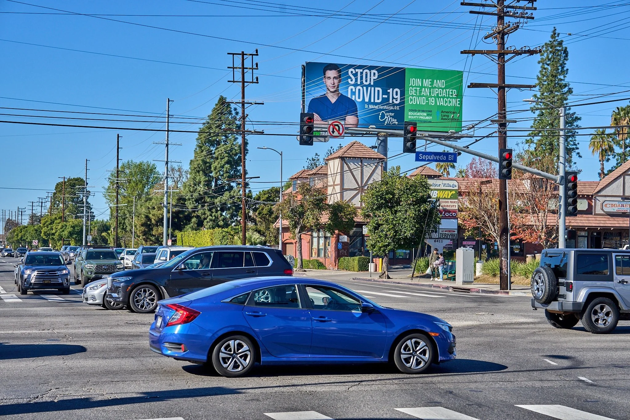 A busy street intersection with several cars, including a blue sedan, and a billboard promoting COVID-19 vaccination. The intersection has traffic lights and power lines, while shops and greenery are visible in the background. The street sign indicat