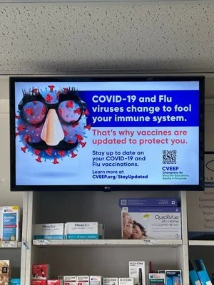 Digital display showing a COVID-19 and flu vaccination advertisement. The ad features an image of a virus with glasses and a nose, alongside text emphasizing the importance of staying updated on vaccinations to protect the immune system. Includes a Q
