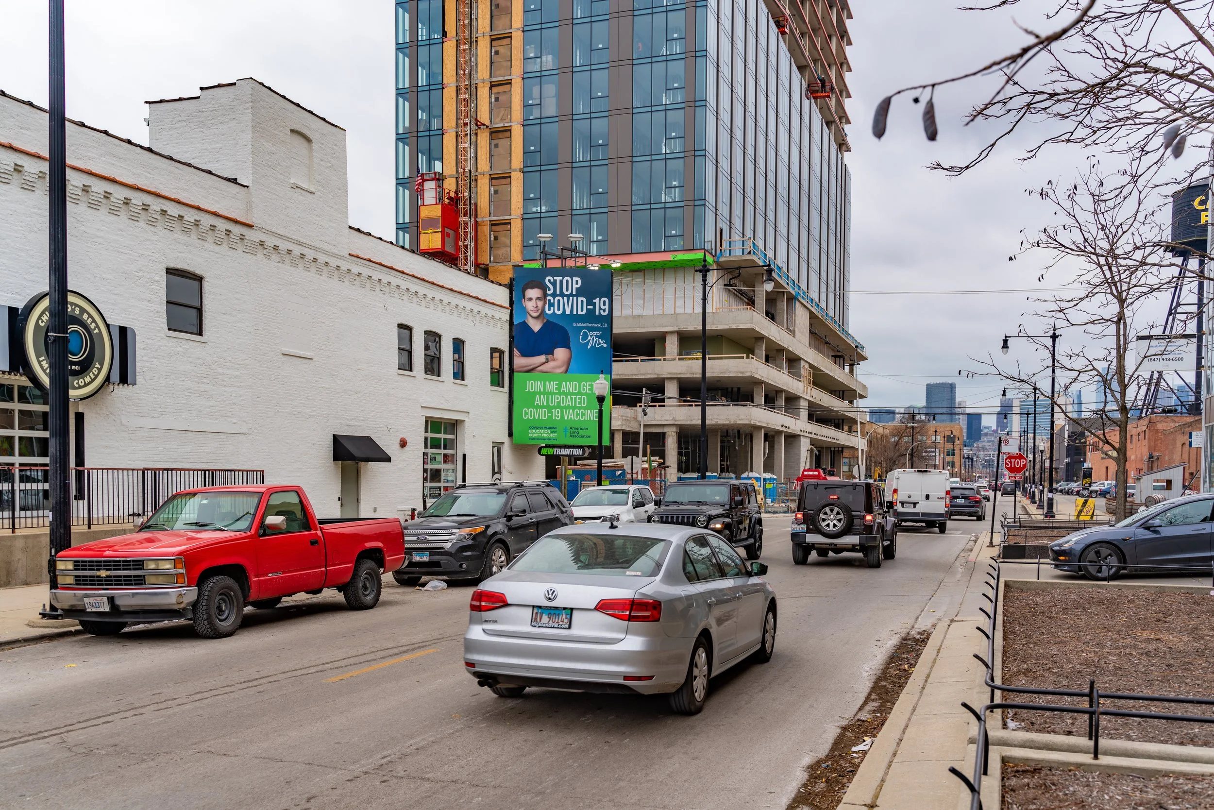 Urban street with traffic, white brick building, under-construction high-rise, and a COVID-19 vaccine billboard.